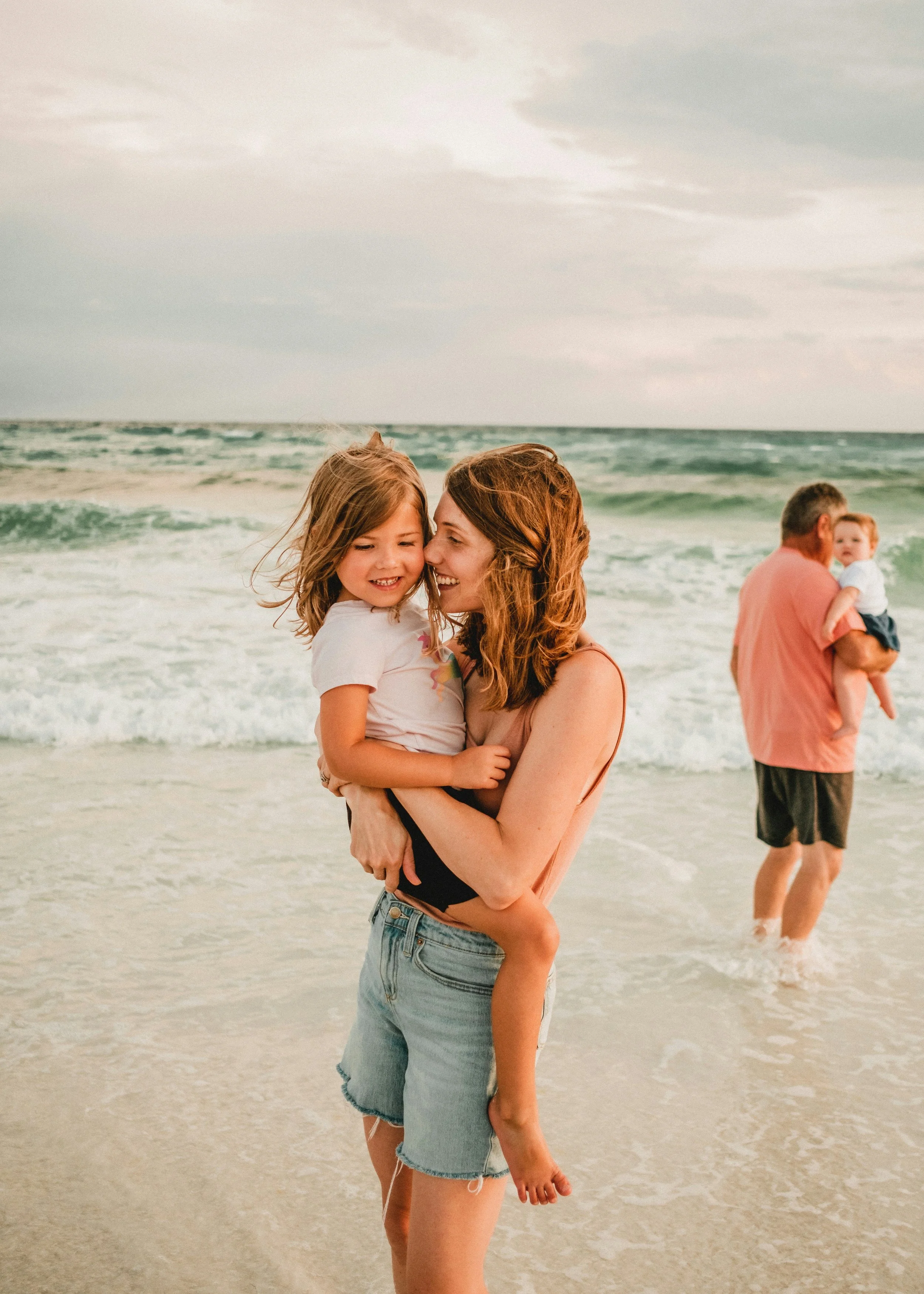 A woman is holding a young girl in her arms on a beach with waves and cloudy sky in the background. A man is in the background holding another child, walking in the shallow water.