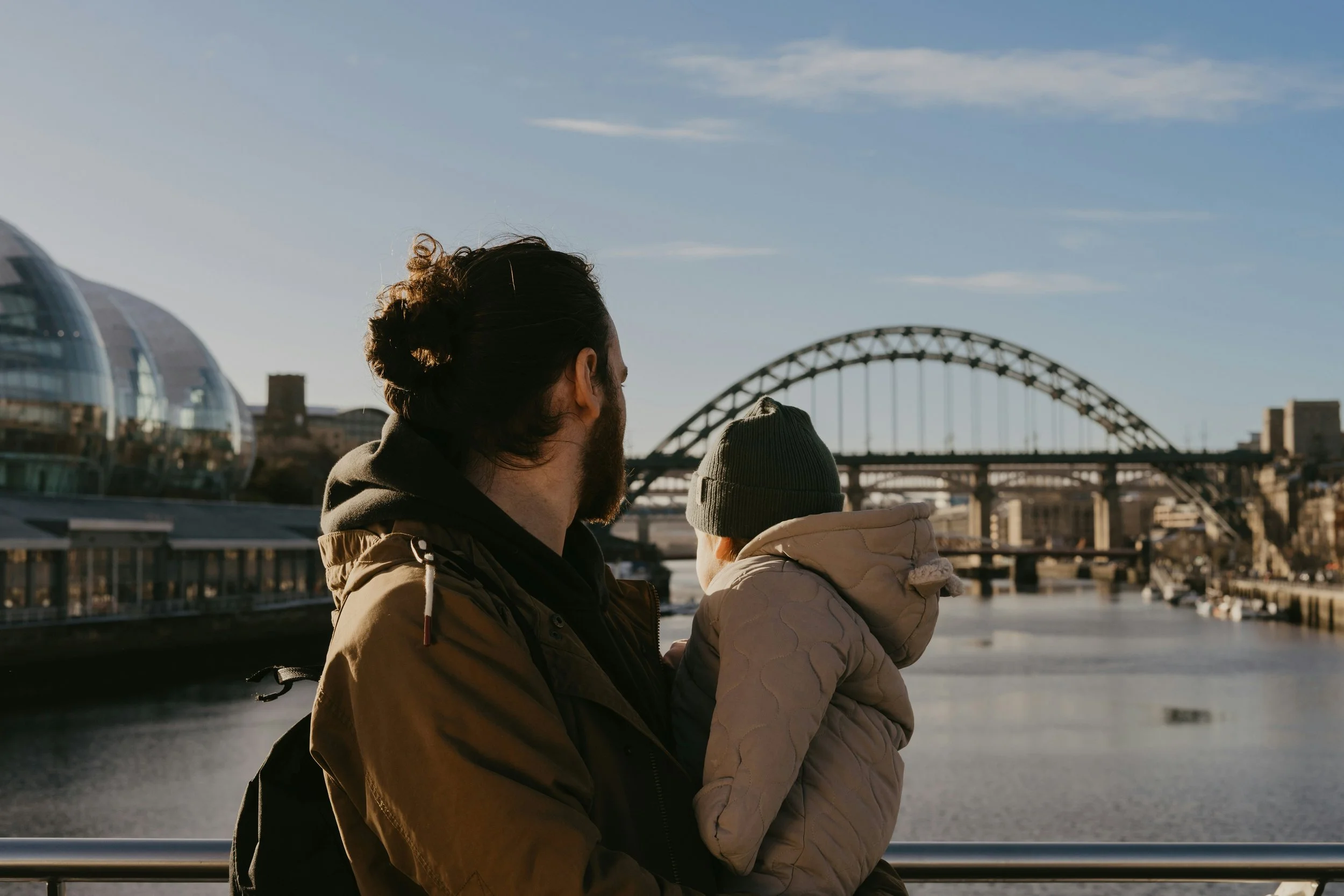 A man and a child view the city skyline and bridge from a riverside promenade on a clear day.