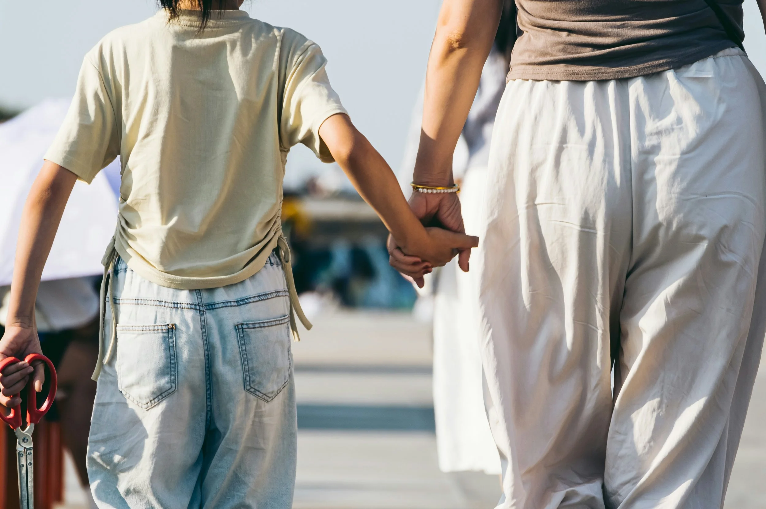 A person wearing a brown shirt and white pants holding hands with a young girl in a beige shirt and denim shorts, with another person holding a pair of red scissors nearby. The scene is outdoors, likely during daytime.