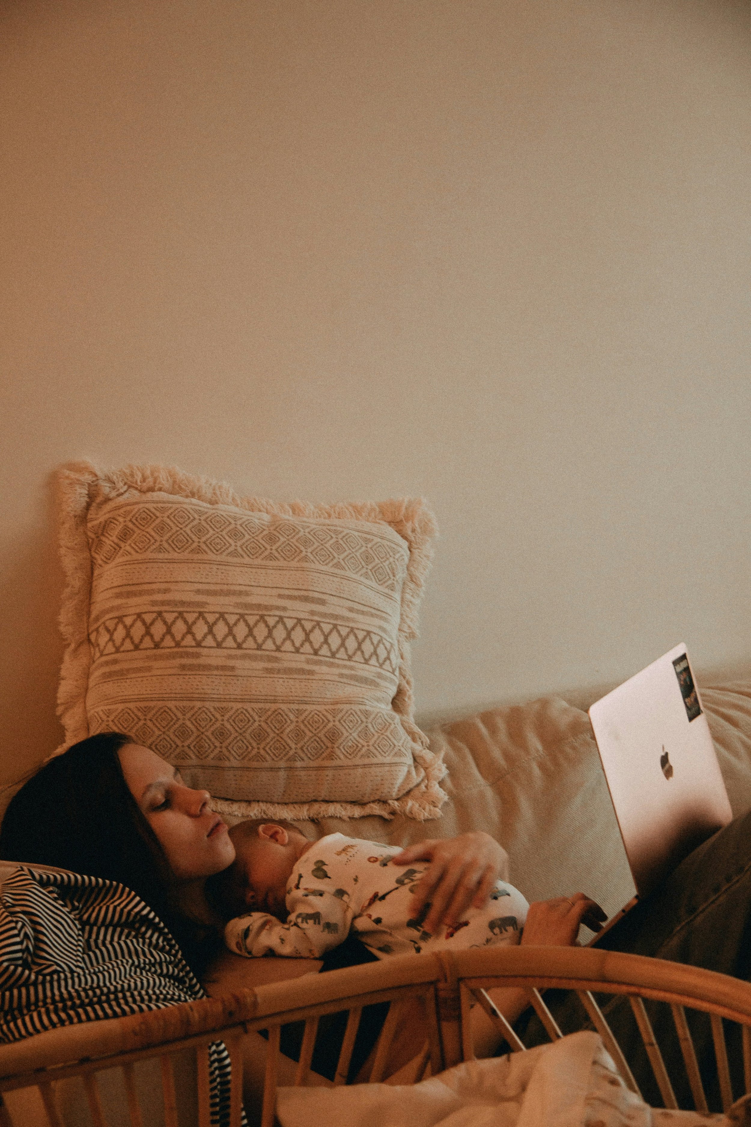 A woman with dark hair and a striped shirt lying on a bed next to a young child with a floral pajama top, both using a laptop.