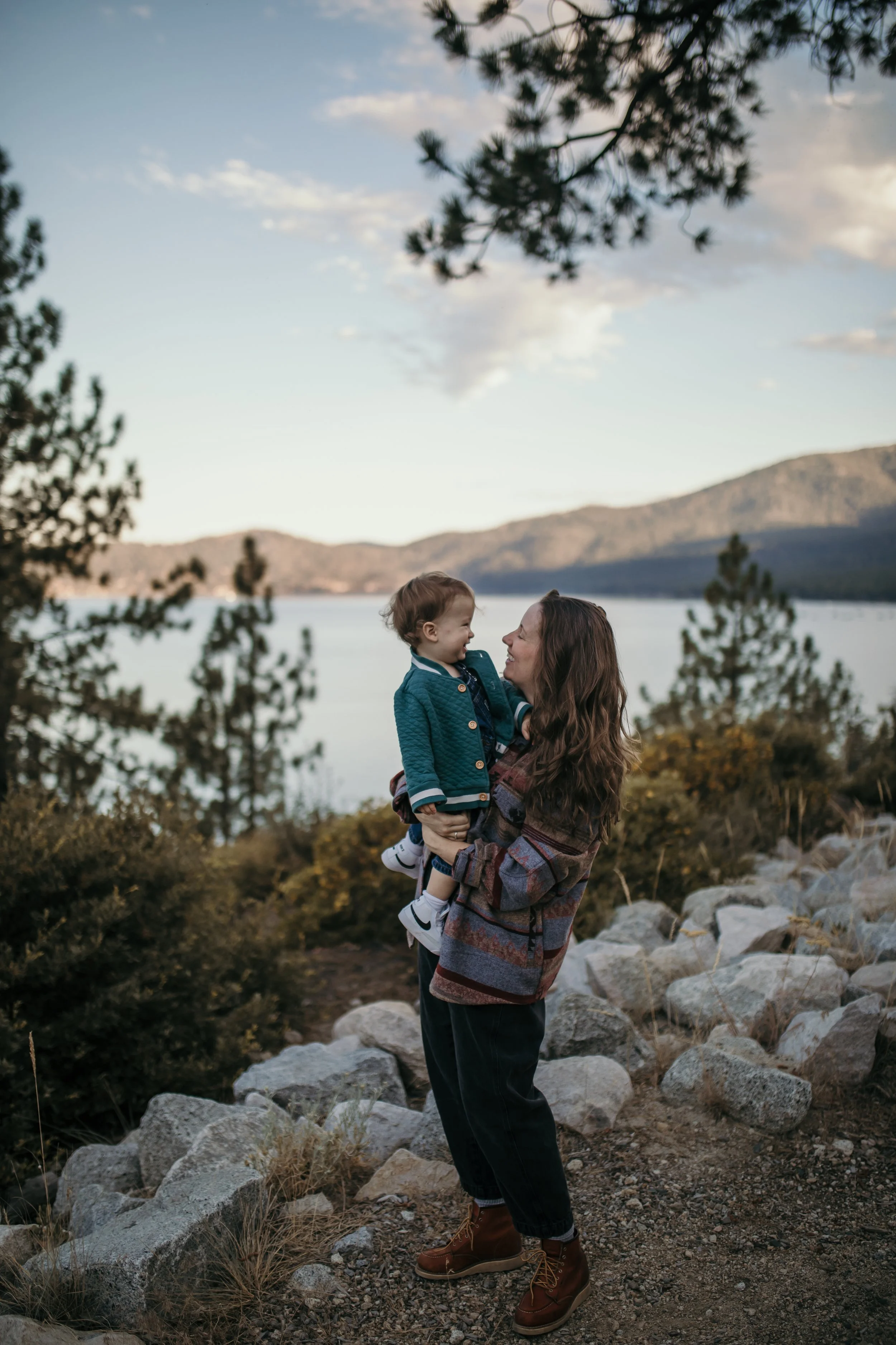 A woman with long brown hair holding a young boy in an outdoor setting near a lake with mountain background during late afternoon or early evening. They are surrounded by rocks and trees, with the woman smiling at the boy.