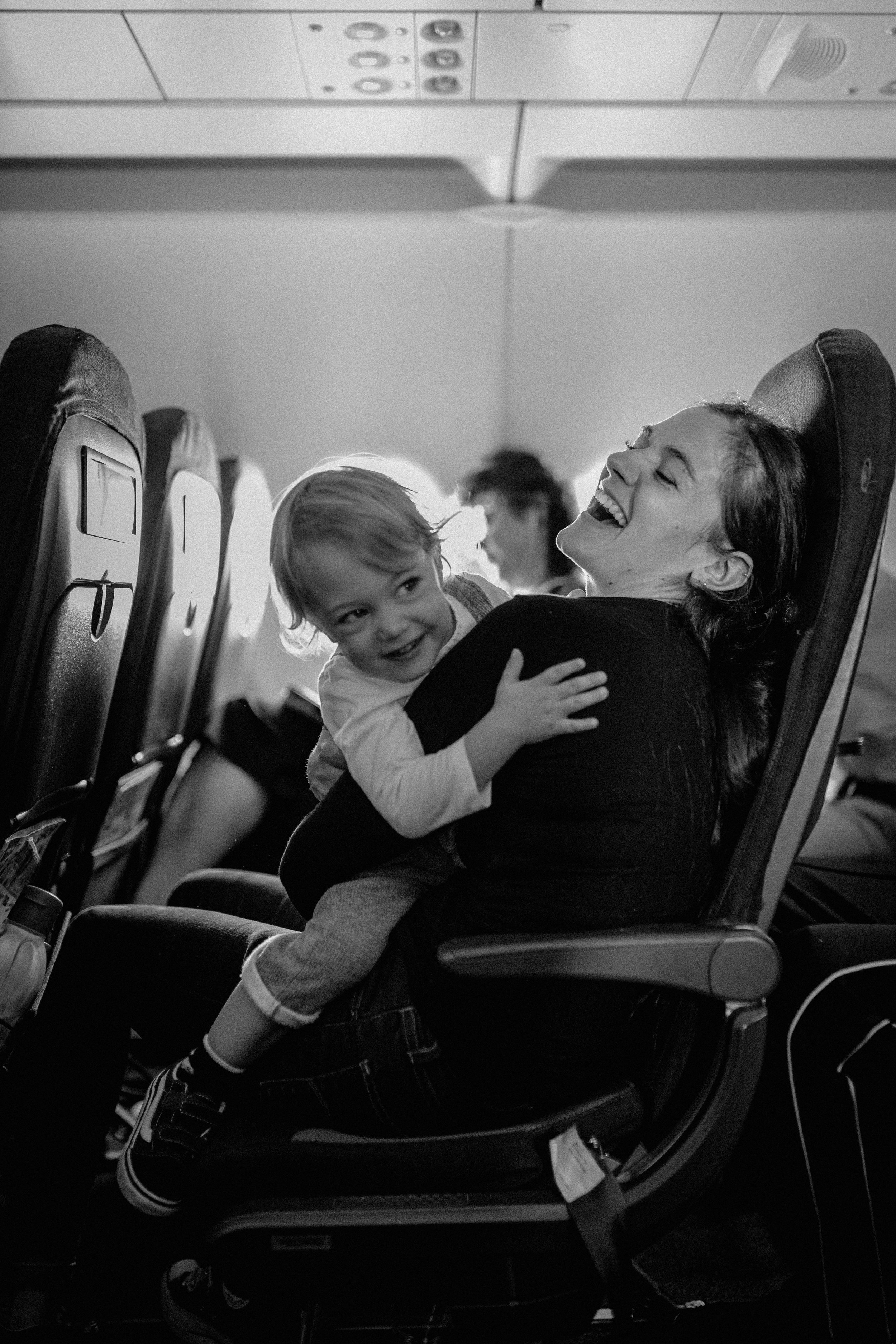 A woman and a young girl sitting on an airplane, laughing and hugging each other. The woman is seated with her head back, eyes closed, and smiling broadly. The girl is looking at the woman with a joyful expression. The airplane seats and overhead compartments are visible in the background.