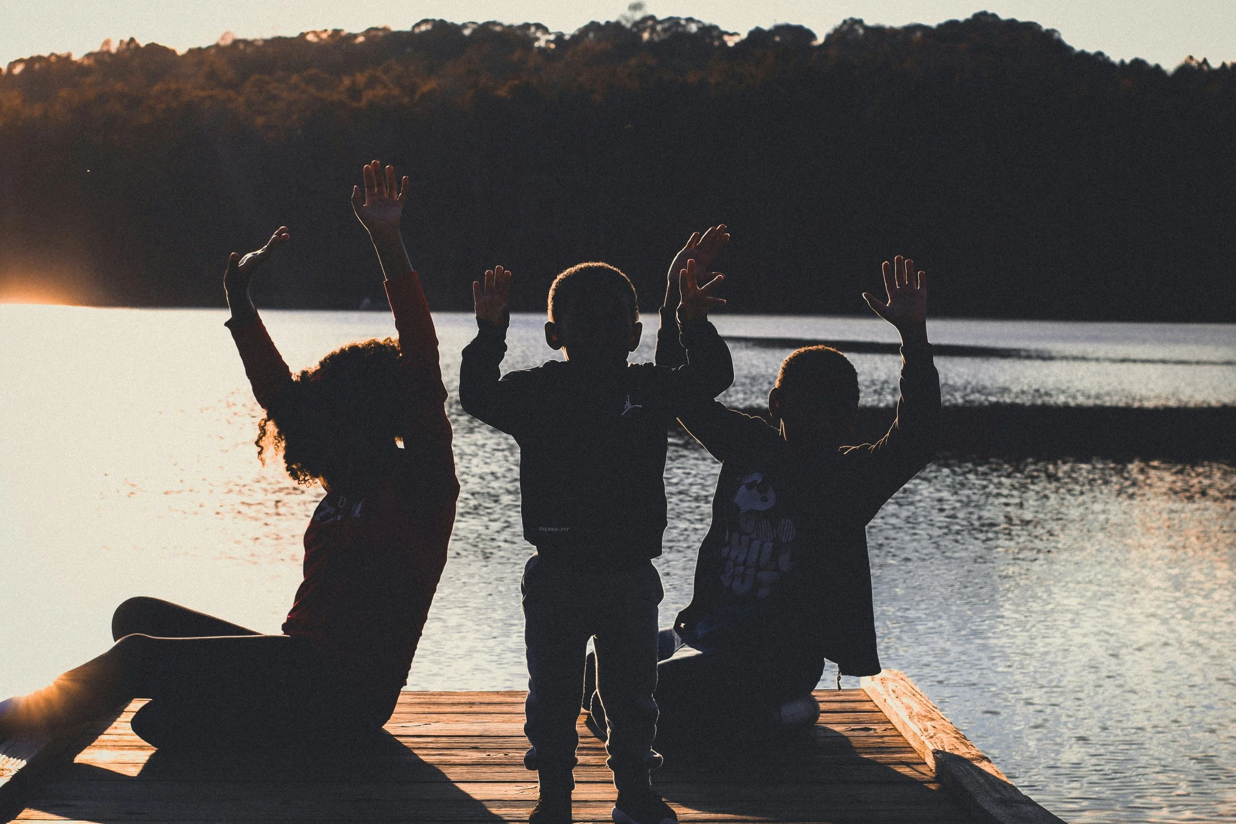 Silhouettes of children and an adult sitting and standing on a wooden dock by a lake at sunset, with a mountain range in the background.