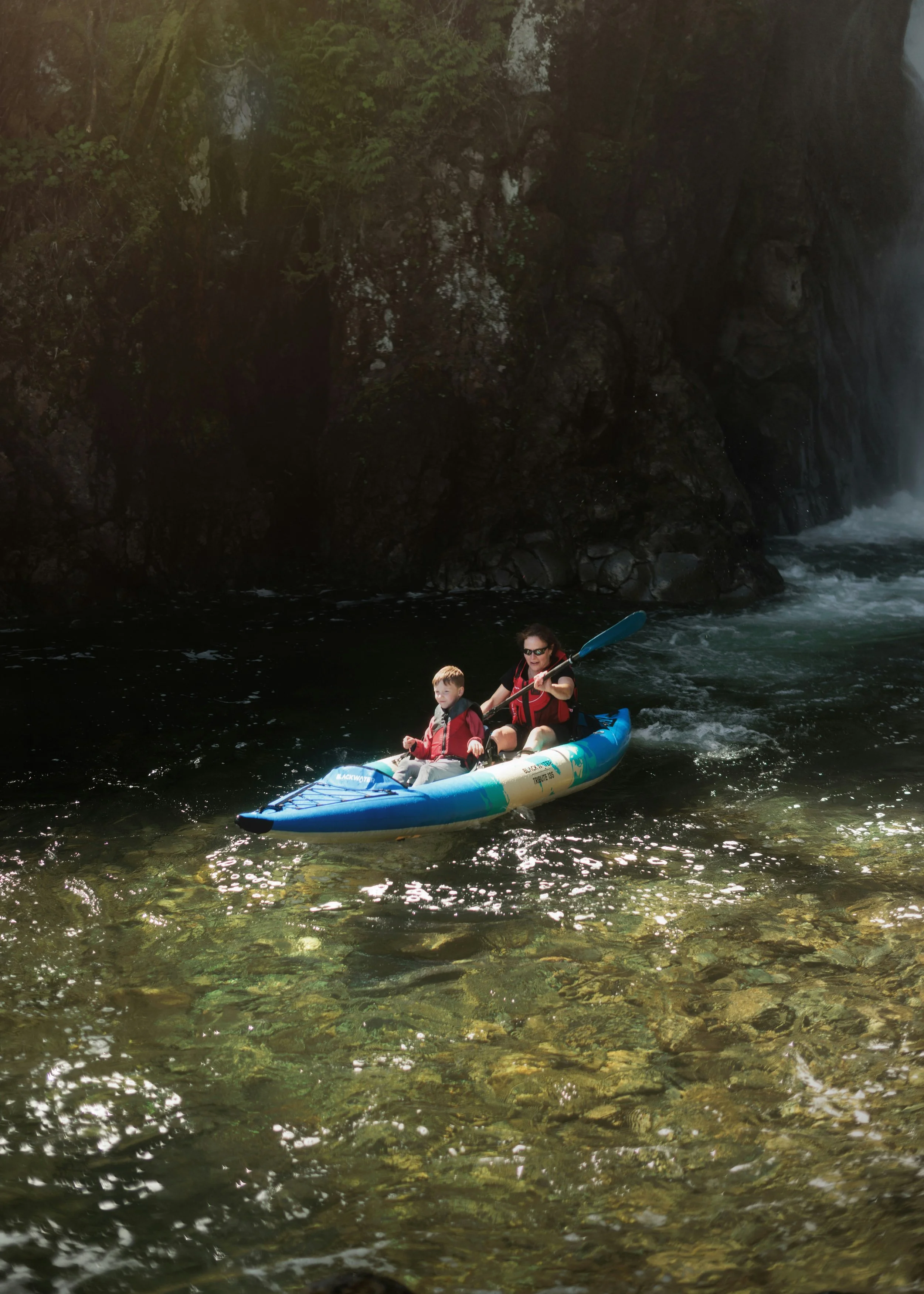 A woman and a young child in a blue kayak paddling on a river with rocky cliffs and green trees in the background.