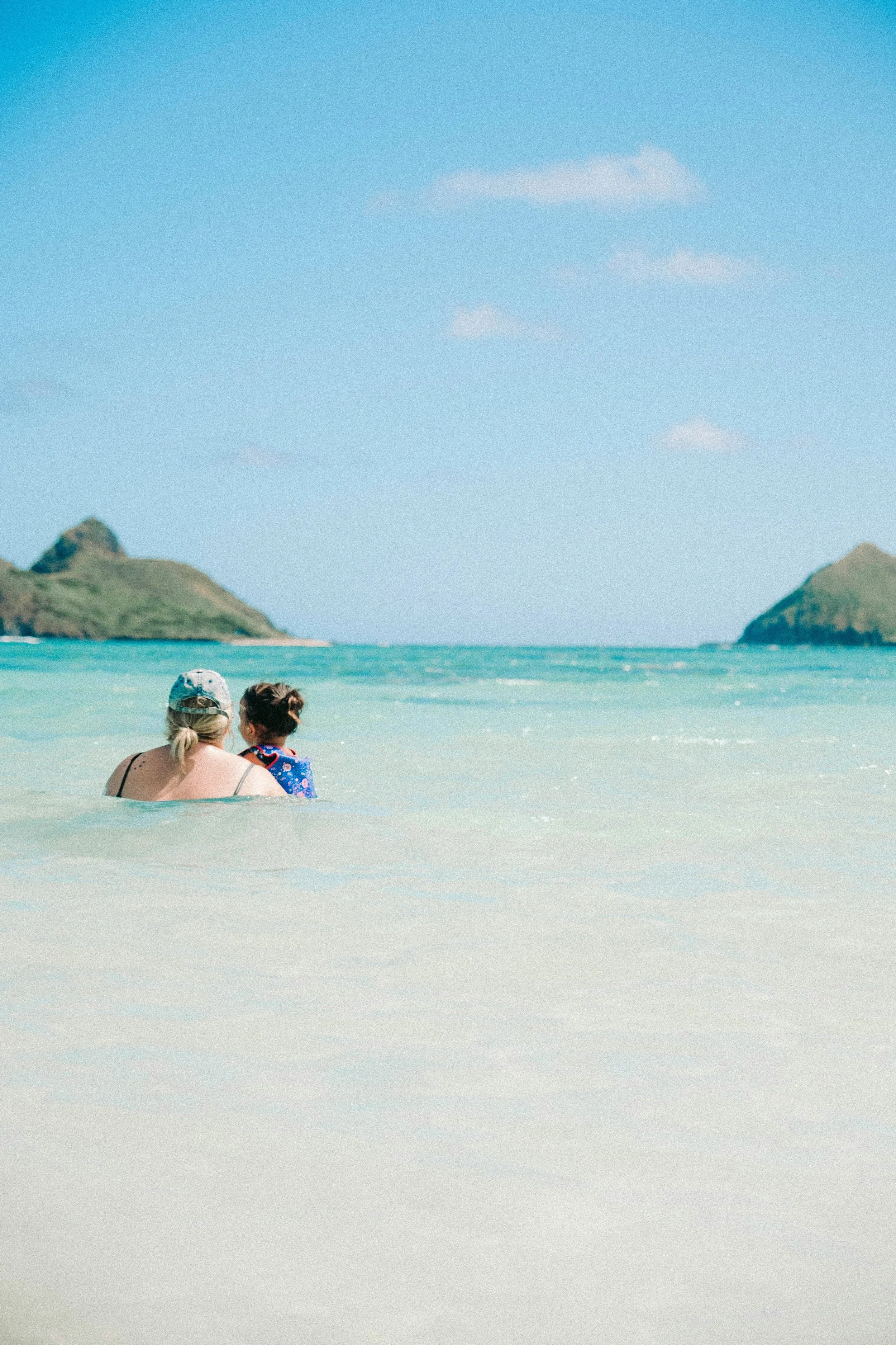Two women sitting in the shallow ocean water, facing away from the camera. One wears a baseball cap and has blonde hair; the other has dark hair in a bun. Two small islands are visible in the distance under a clear blue sky.