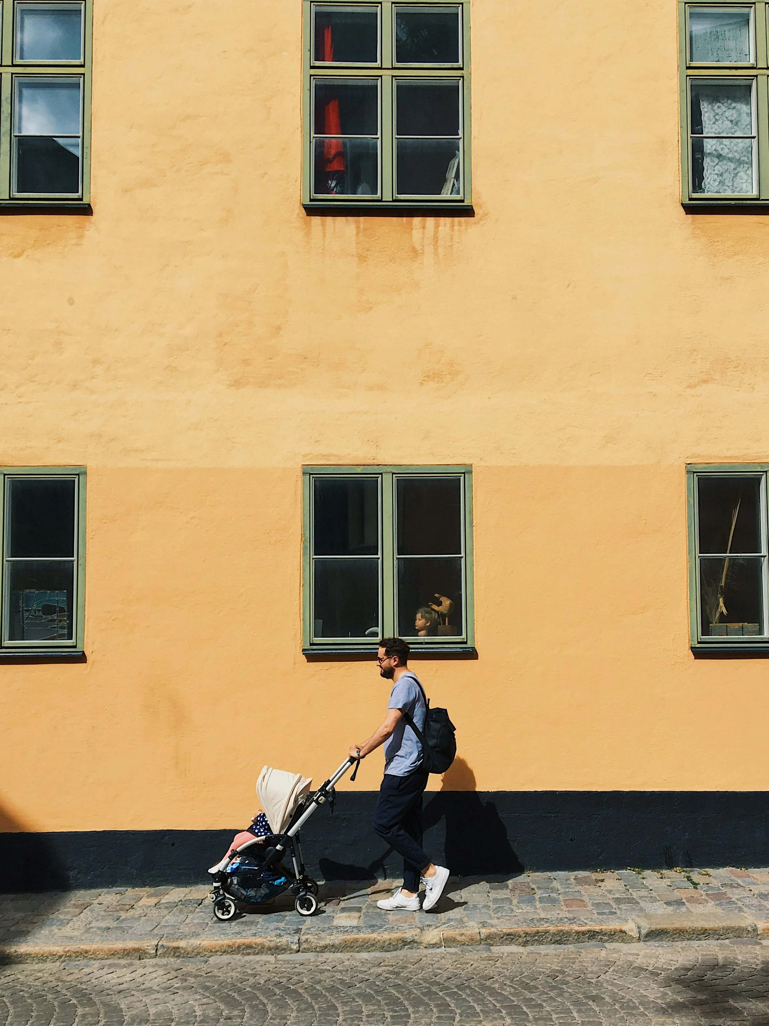 A man pushing a baby stroller on a cobblestone sidewalk in front of a yellow building with green-framed windows, some of which have curtains or objects inside.