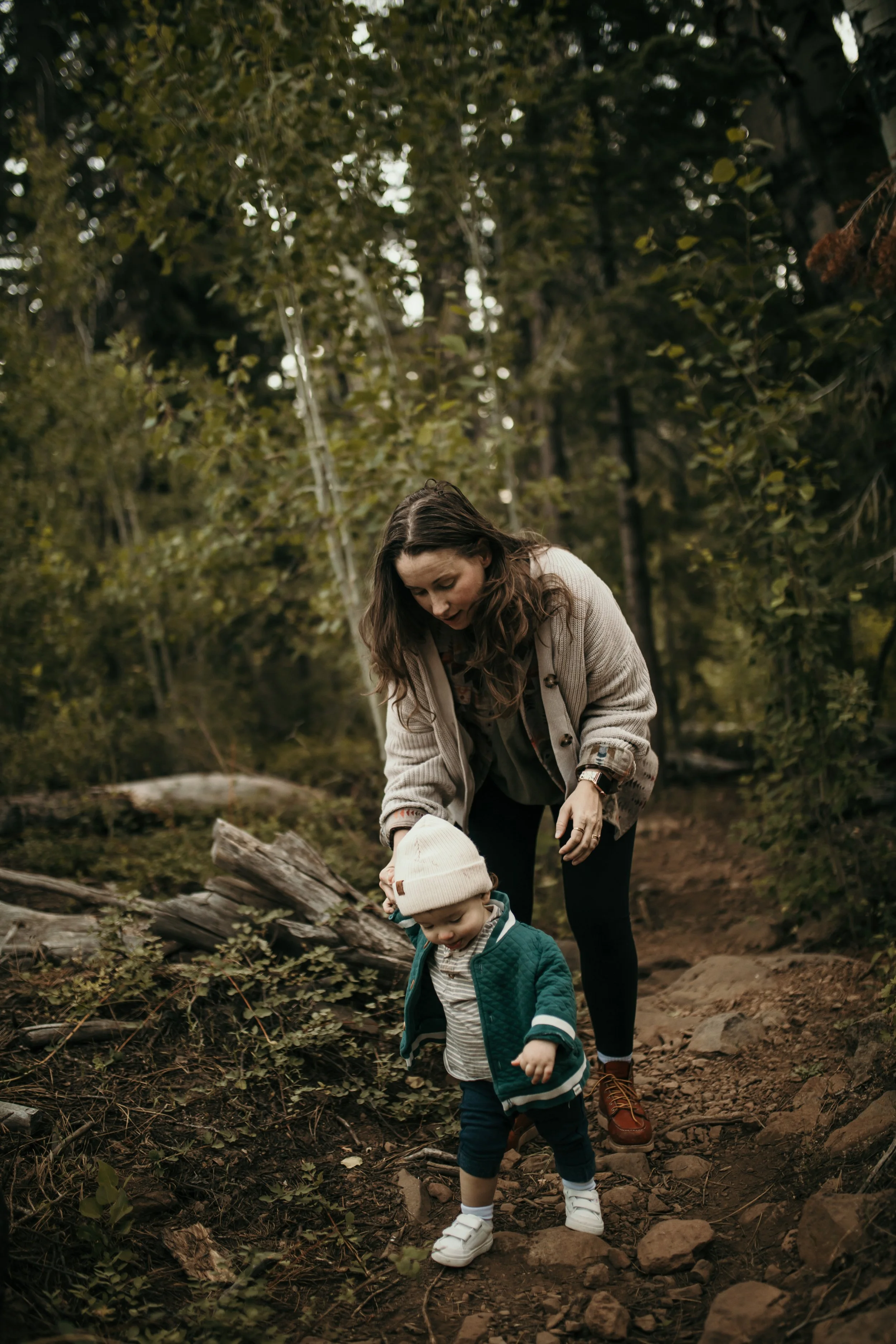 A woman and child exploring a forest trail with fallen logs and scattered rocks, surrounded by dense green foliage.