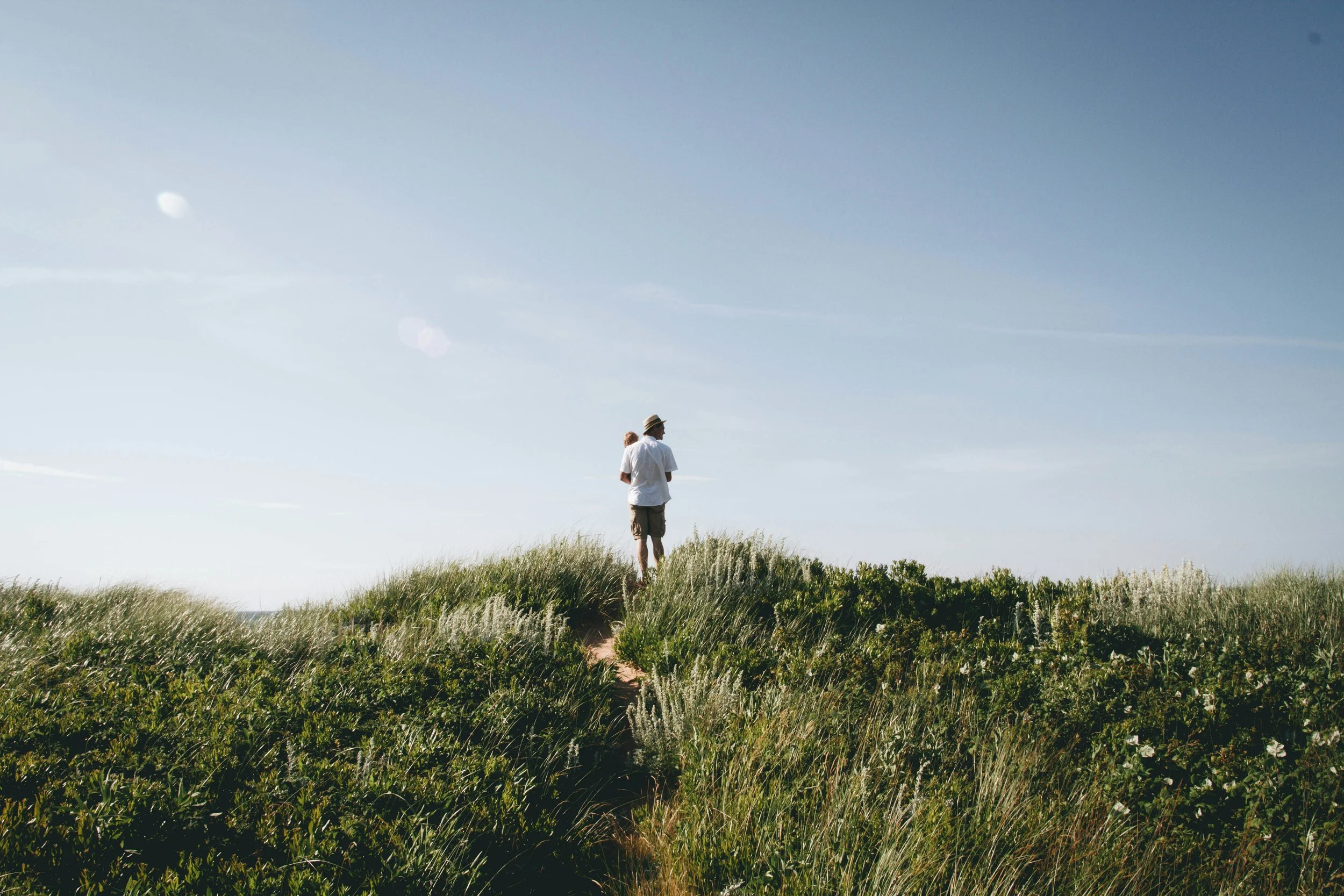 A person wearing a hat and white shirt standing on a grassy hill under a blue sky with a few clouds.