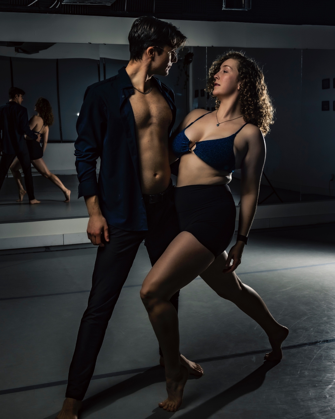 Two dancers practicing in a dance studio, with a mirror reflecting them and other dancers in the background.