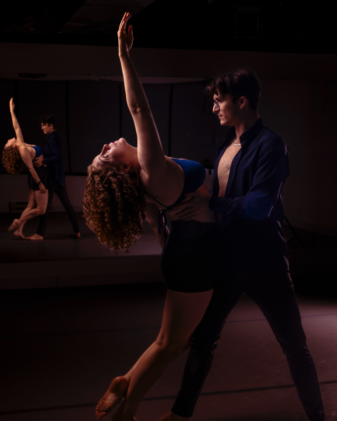A dance rehearsal showing a male dancer holding a female dancer who is leaning back, with other dancers practicing in the background.