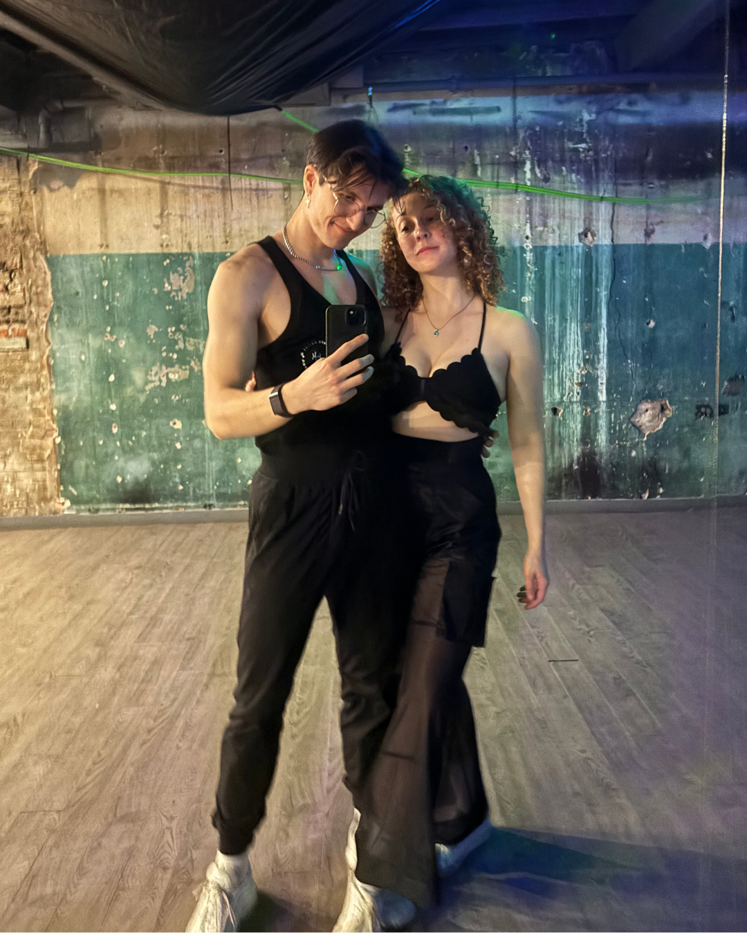 Two young adults taking a mirror selfie in a dance studio with distressed green and exposed brick walls.