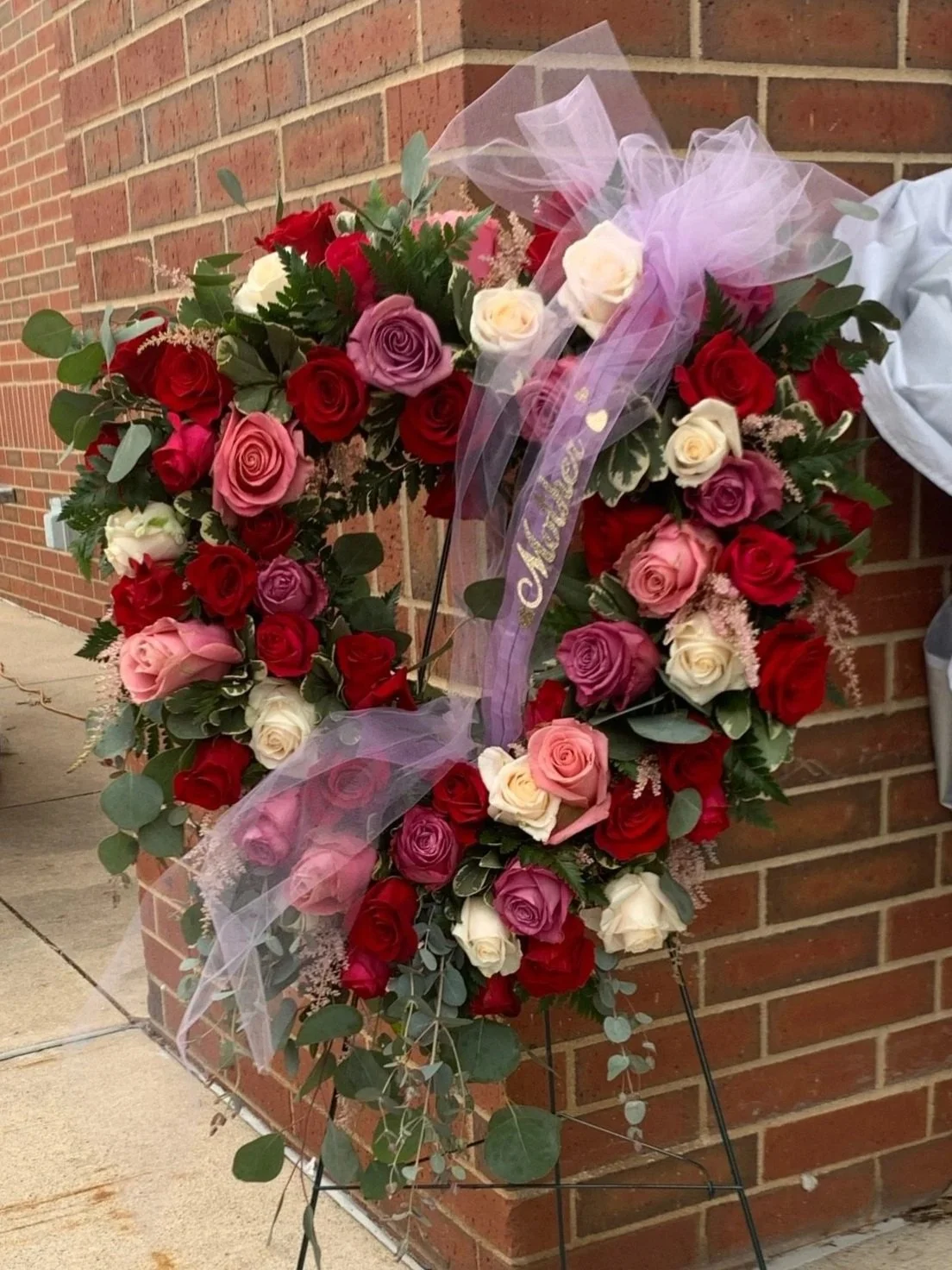 A floral memorial wreath with red, pink, white, and purple roses, decorated with a pink ribbon and mesh fabric, placed against a brick wall.