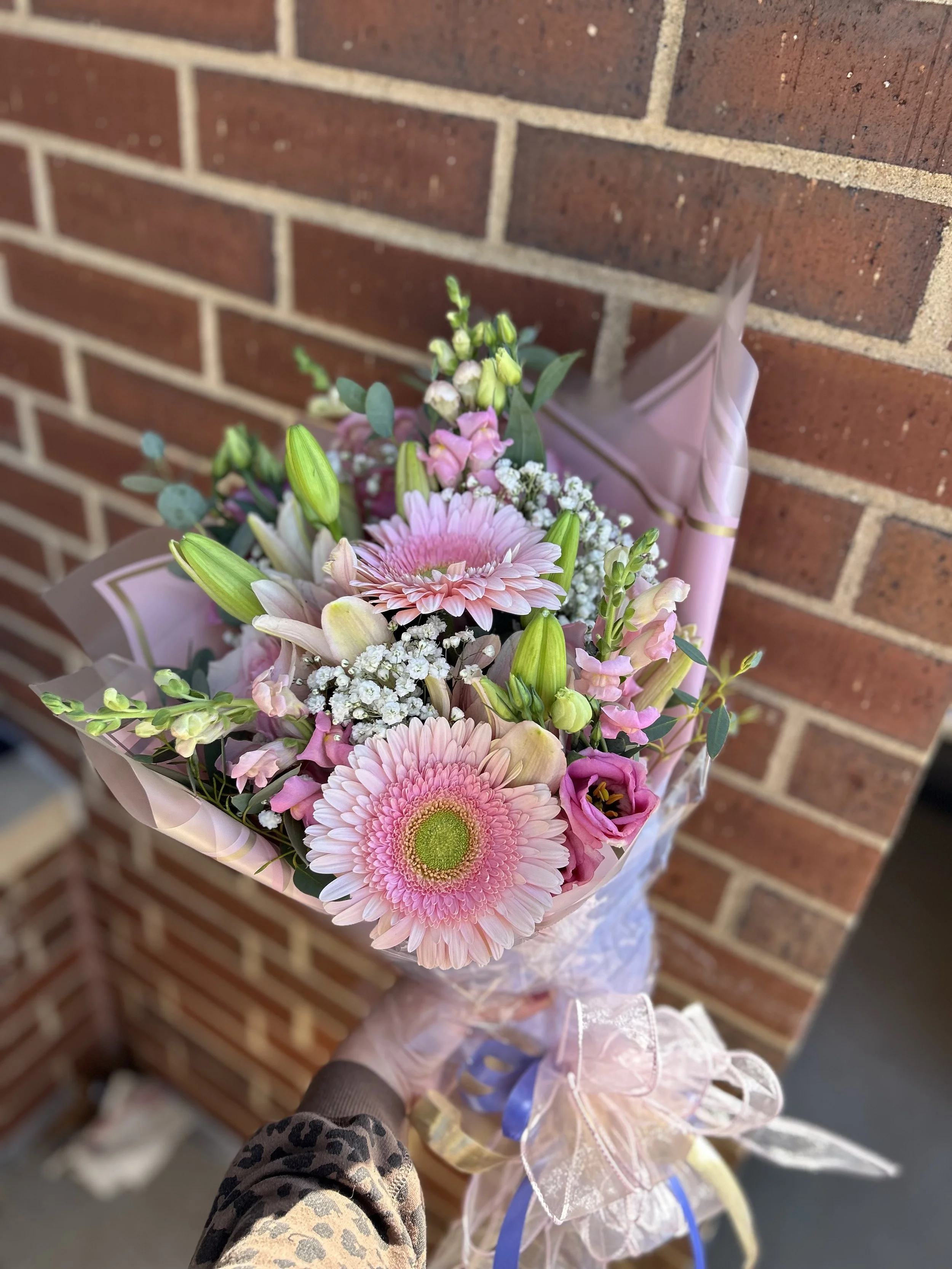 A hand holding a bouquet of pink and white flowers, including Gerbera daisies, lilies, and small filler flowers, wrapped in pink tissue paper with bows, against a brick wall background.