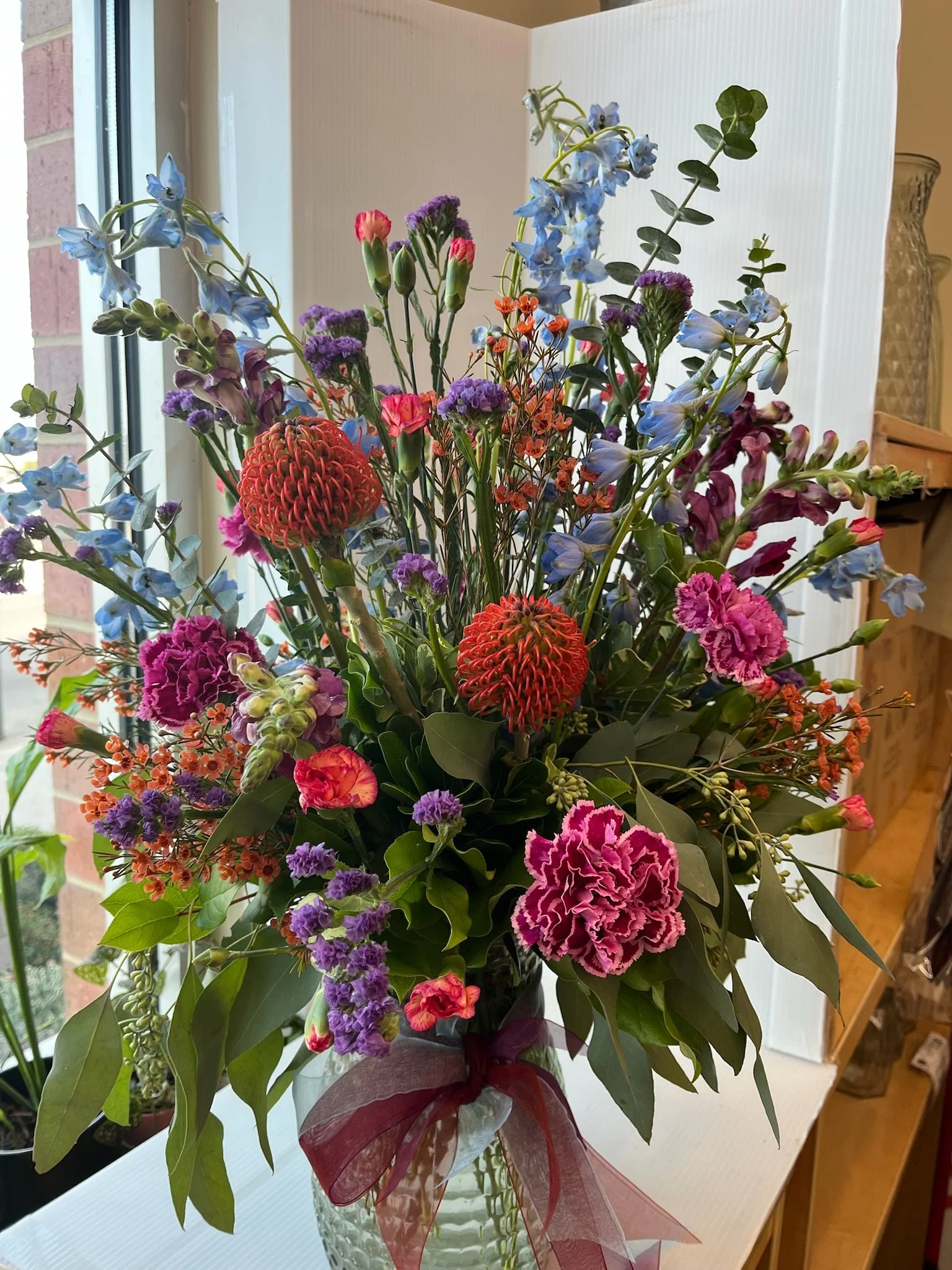 A colorful bouquet of various flowers in a glass vase with a ribbon, placed on a white surface by a window.