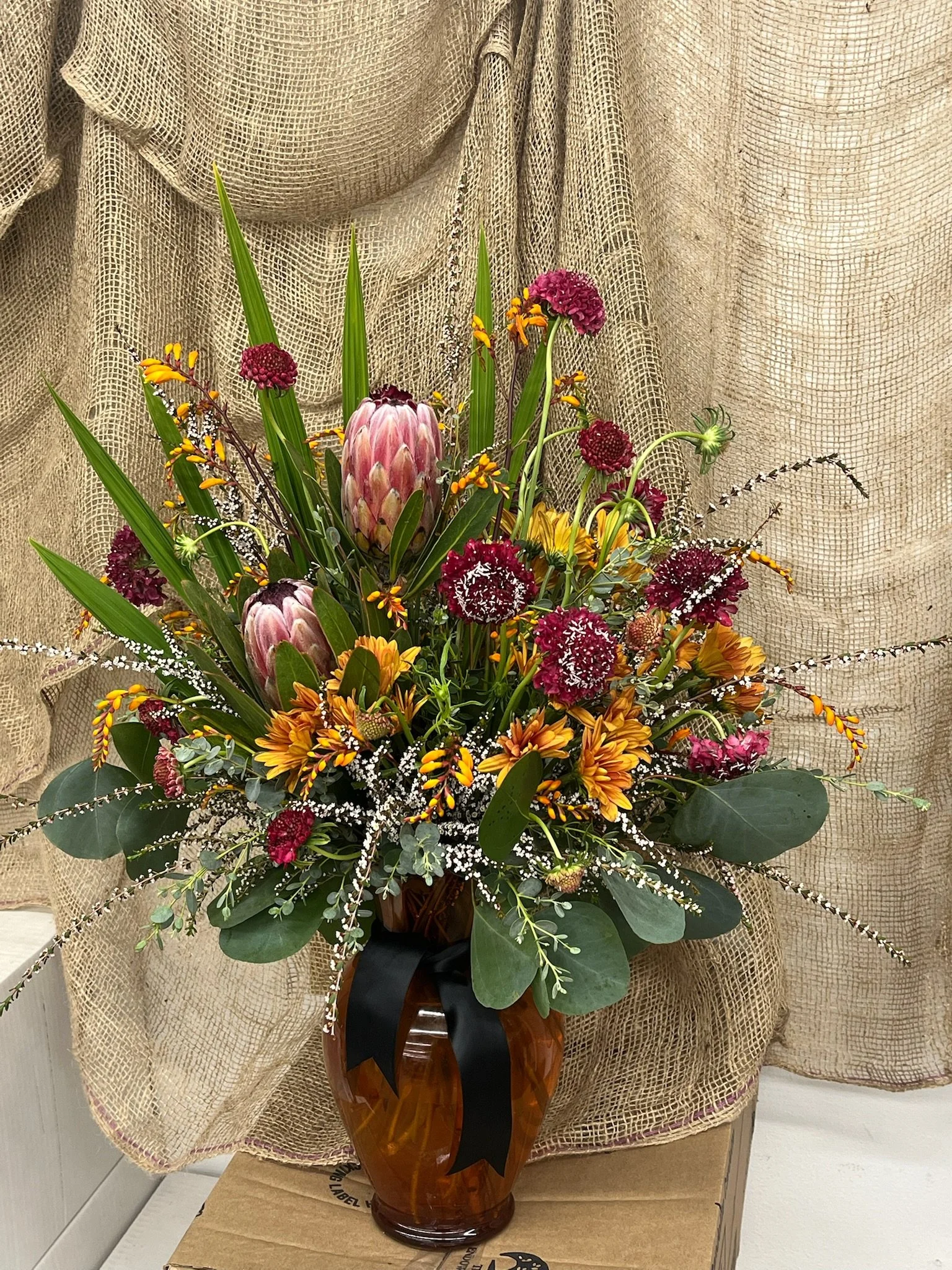 A colorful flower arrangement in a brown vase, featuring protea, maroon scabiosa, yellow daisies, orange and yellow chrysanthemums, white baby's breath, and green foliage, set against a textured beige backdrop.