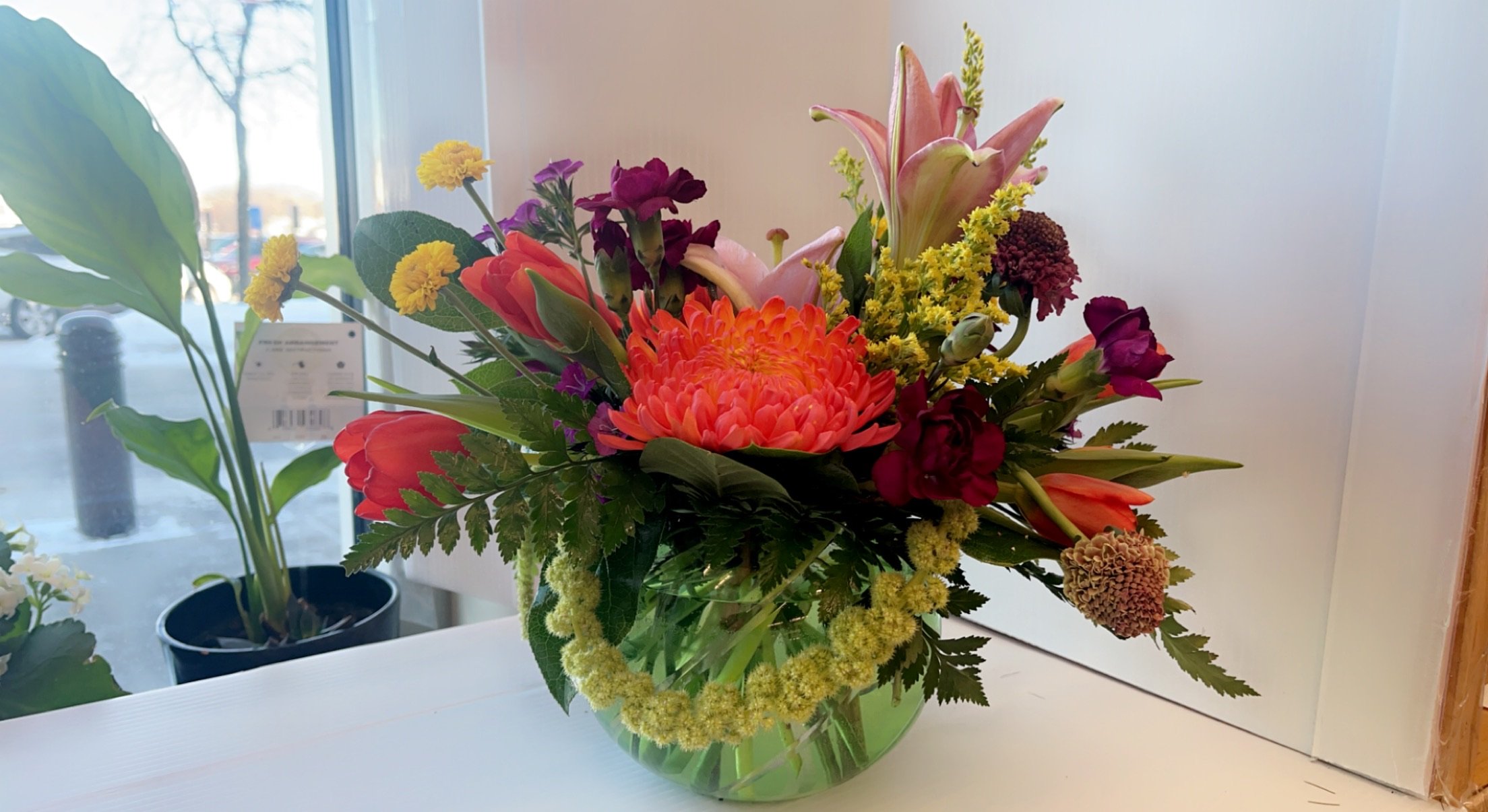 Colorful mixed flower bouquet in a clear glass vase placed on a white surface near a window, featuring pink, orange, yellow, and purple flowers with green foliage.