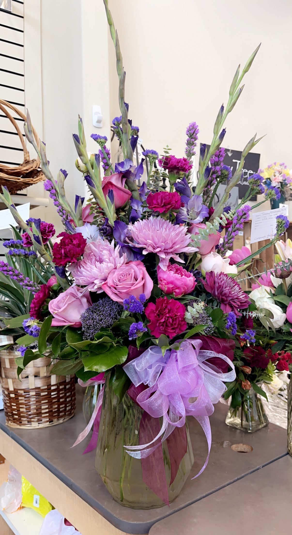 Pink, purple, and white mixed flower bouquet in a glass vase with ribbons, including roses, chrysanthemums, and delphiniums.