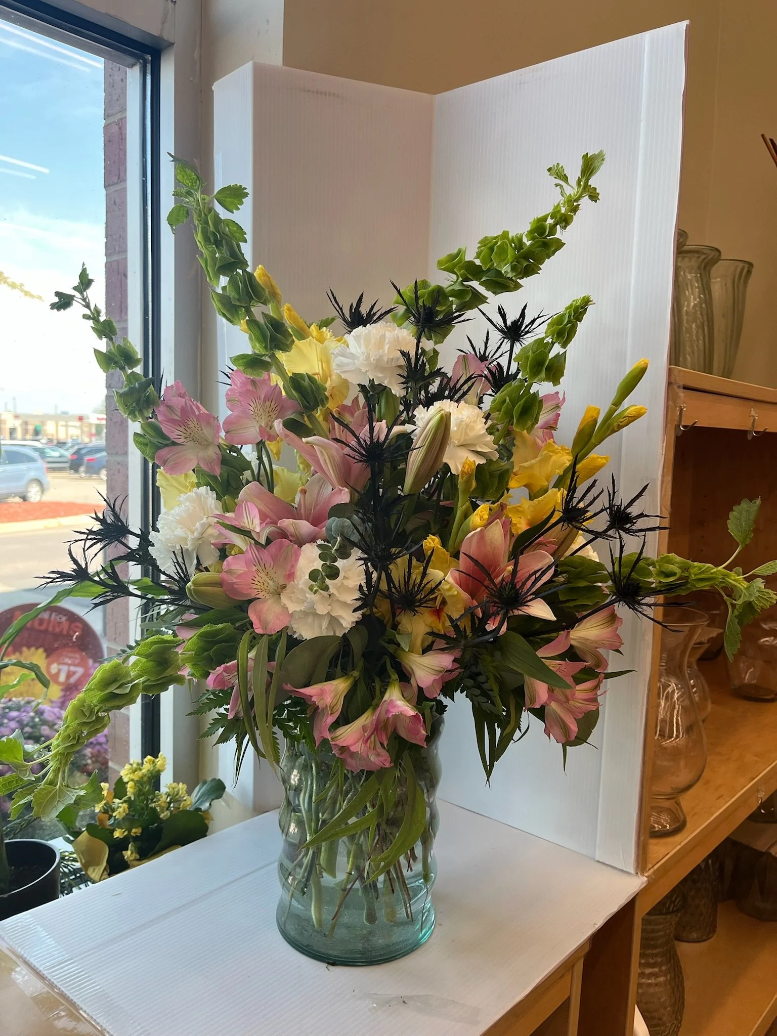 A glass vase filled with a bouquet of pink, white, yellow, and green flowers on a white surface near a window with a view of parked cars outside.