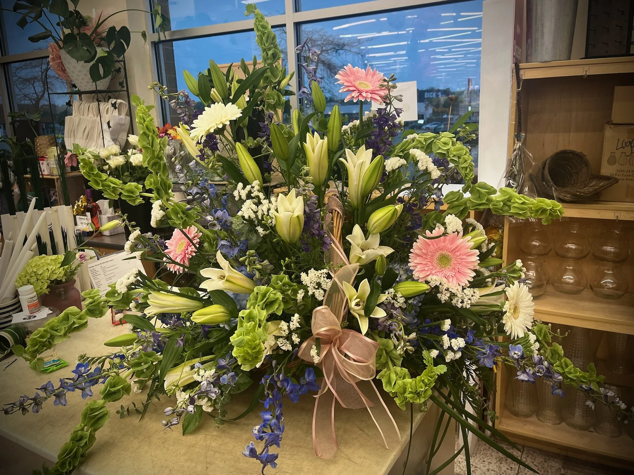 A large, colorful flower arrangement with white lilies, pink and white gerbera daisies, white carnations, purple delphiniums, and green foliage, decorated with a pink ribbon, placed on a counter inside a flower shop.