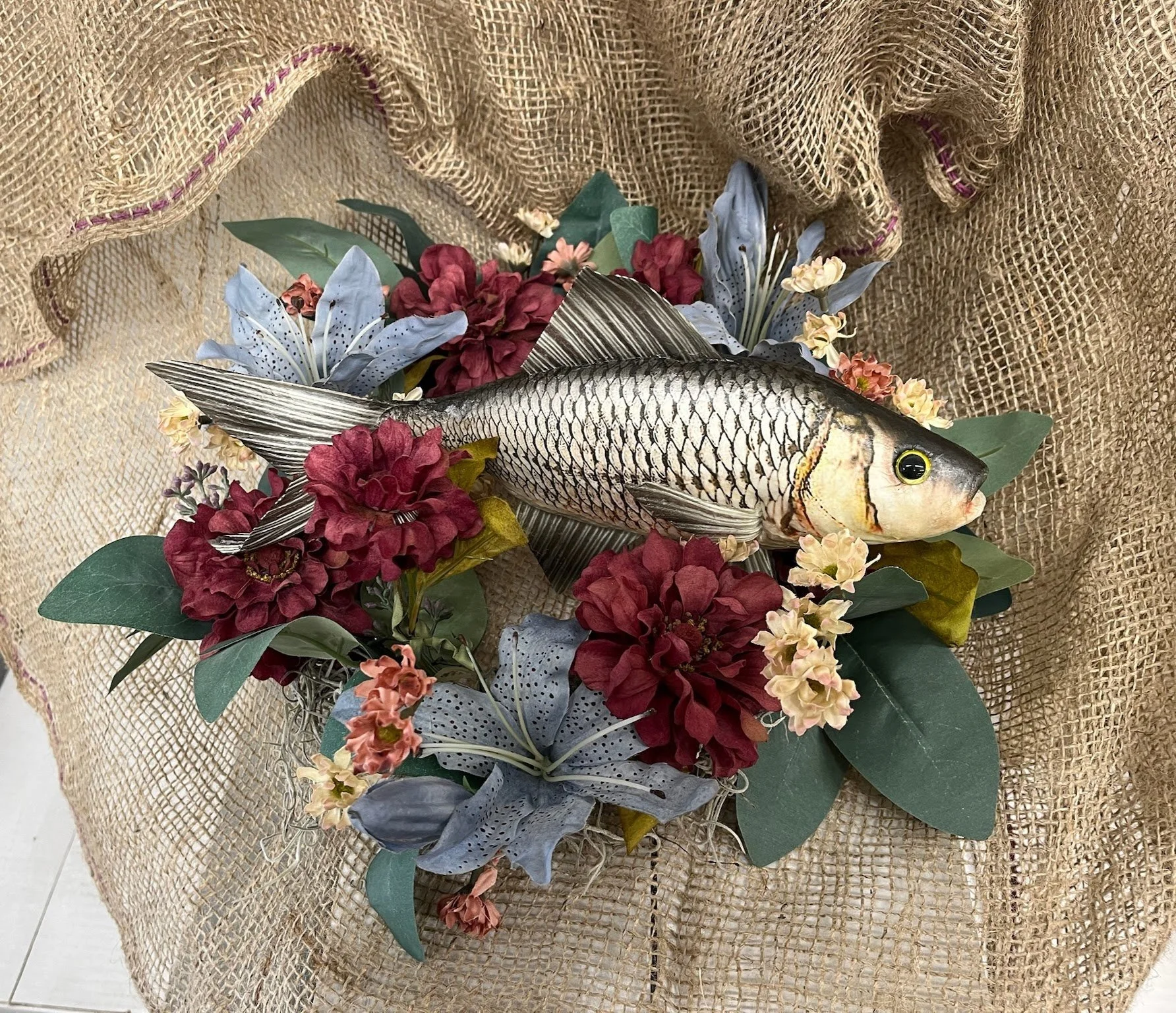 A fish surrounded by colorful artificial flowers and greenery, placed on a burlap fabric background.