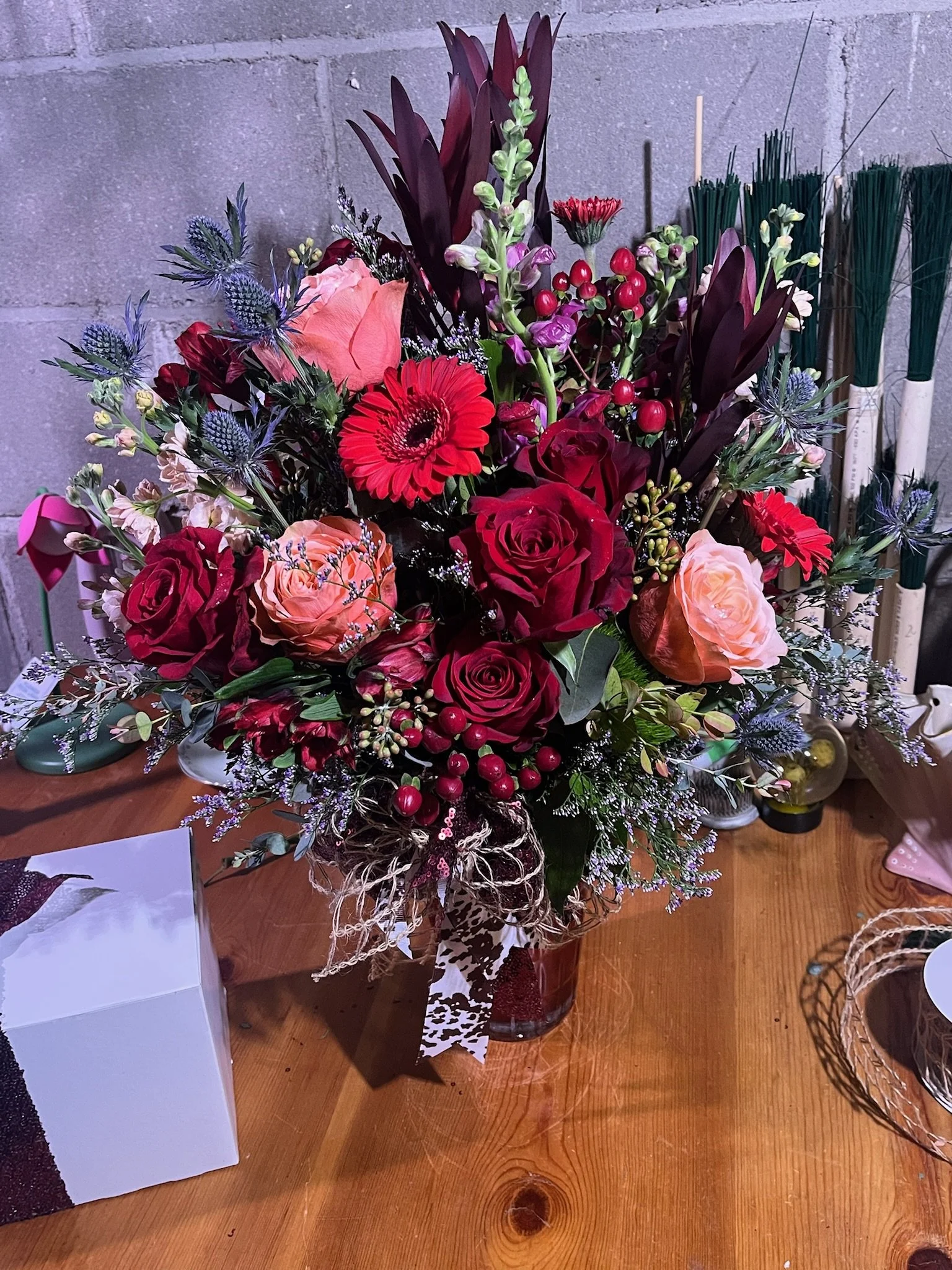 A colorful bouquet of flowers in a glass vase sitting on a wooden table. The arrangement includes roses, gerbera daisies, berries, and various greenery.