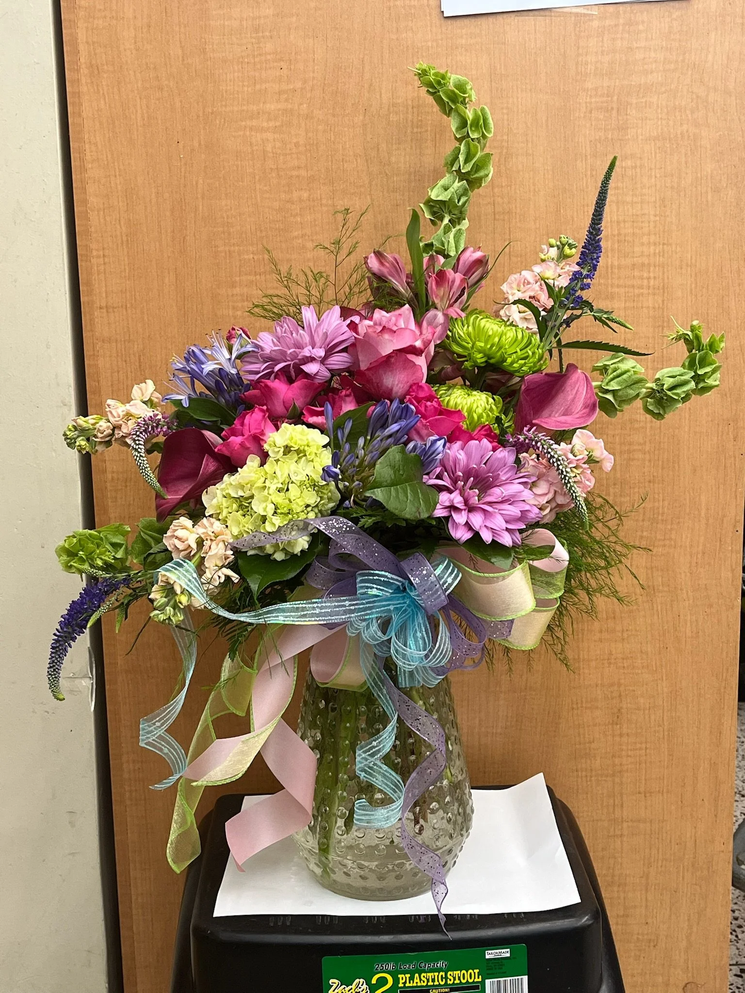 A colorful floral bouquet in a textured glass vase with pastel ribbons, placed on a black plastic stool in front of a wooden background.