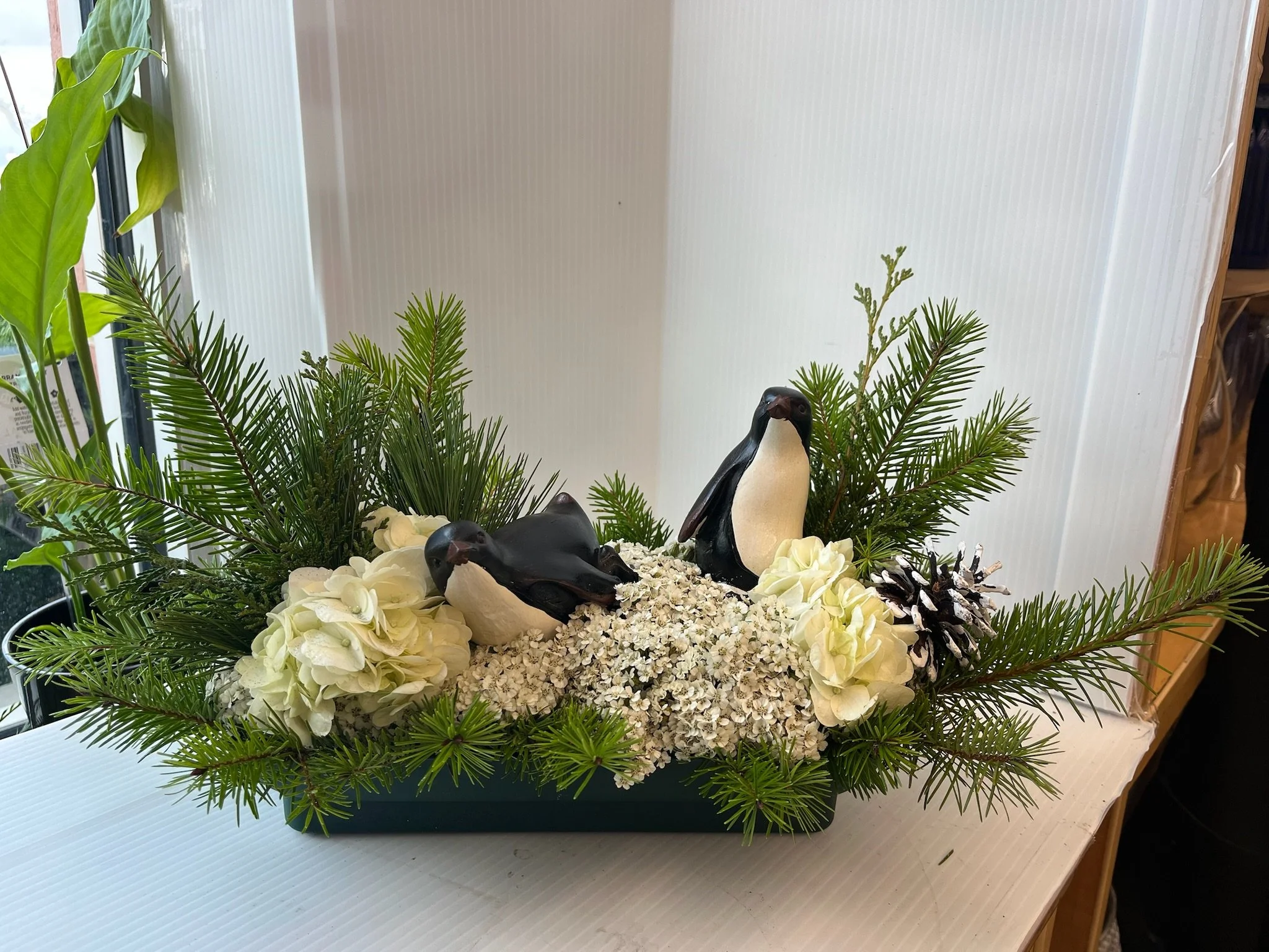 Decorative arrangement of white hydrangeas, white flowers, pine branches, pinecones, and two black and white penguin figurines in a blue container.