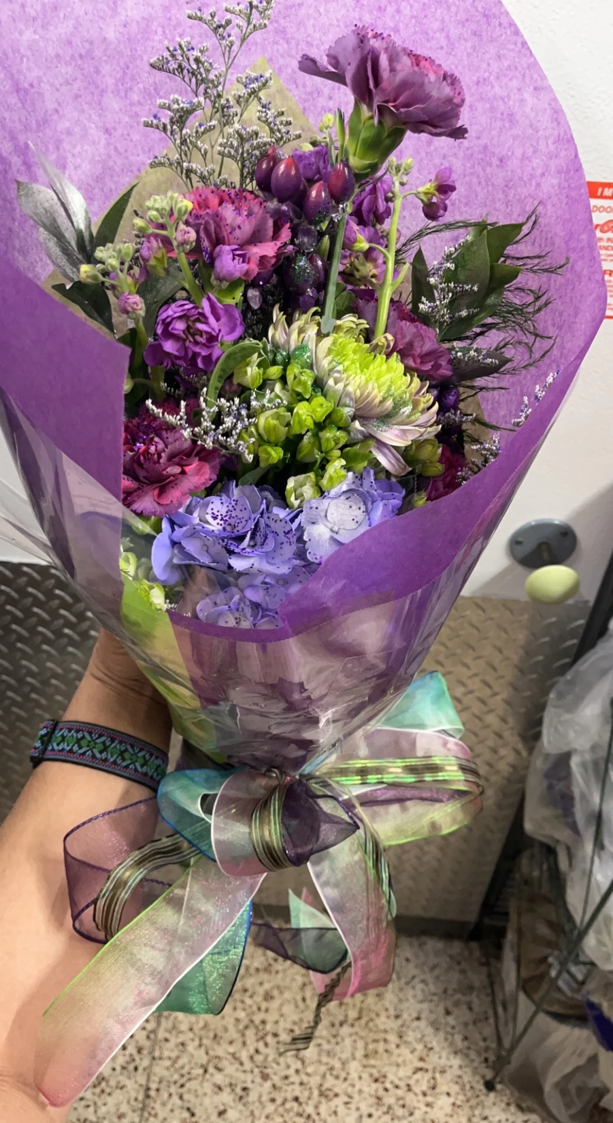 Colorful bouquet of various purple and pink flowers wrapped in purple and transparent paper, tied with a multicolored ribbon.