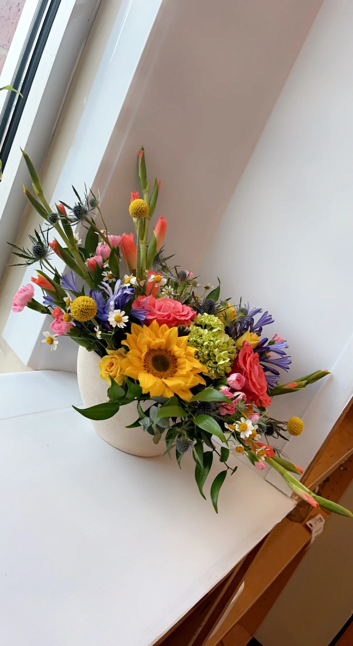 Colorful flower arrangement with sunflowers, roses, daisies, and other mixed flowers in a white vase on a white table near a window.