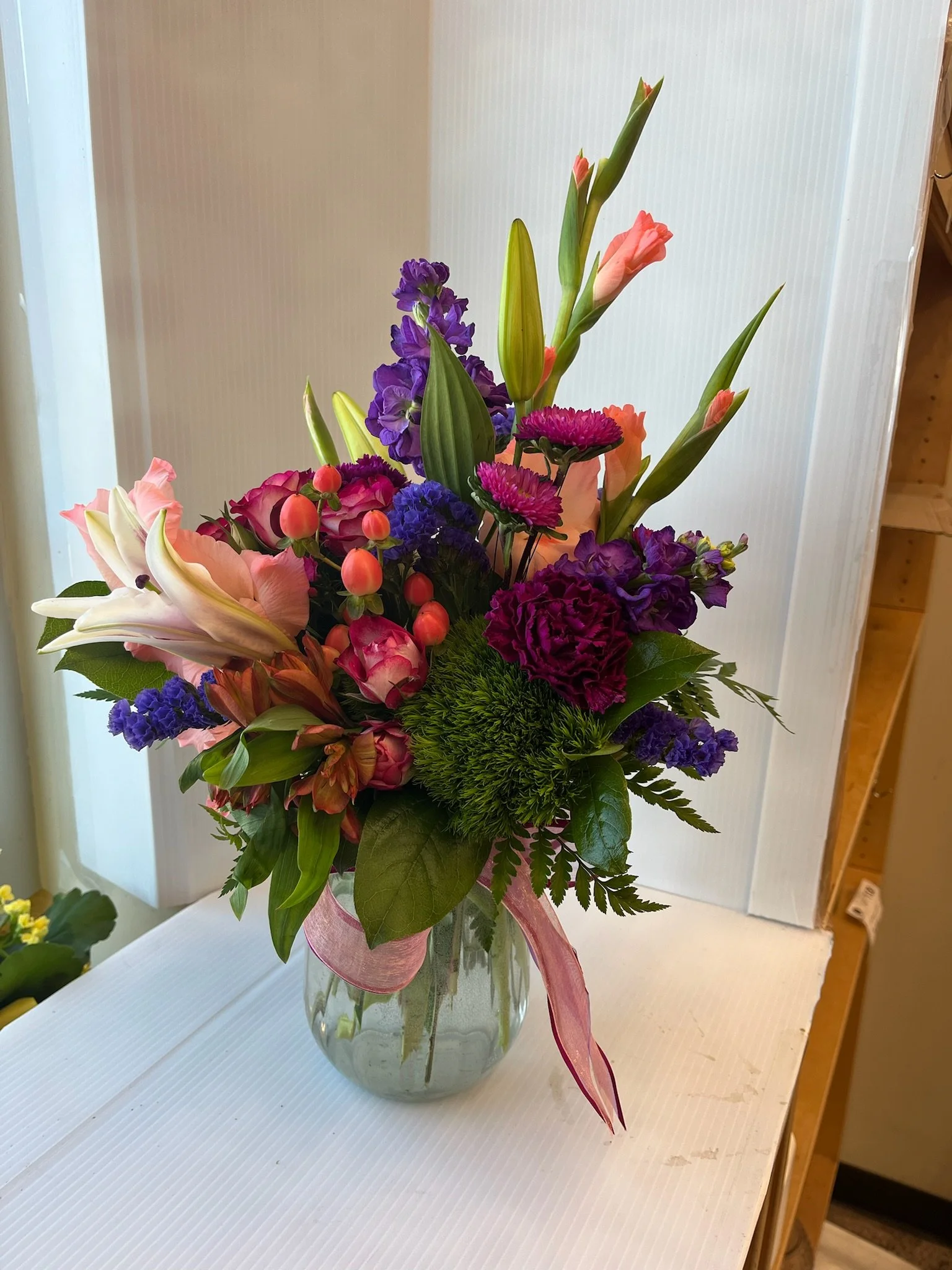 Colorful flower bouquet with pink lilies, purple irises, pink roses, and greenery in a clear glass vase on a white surface.