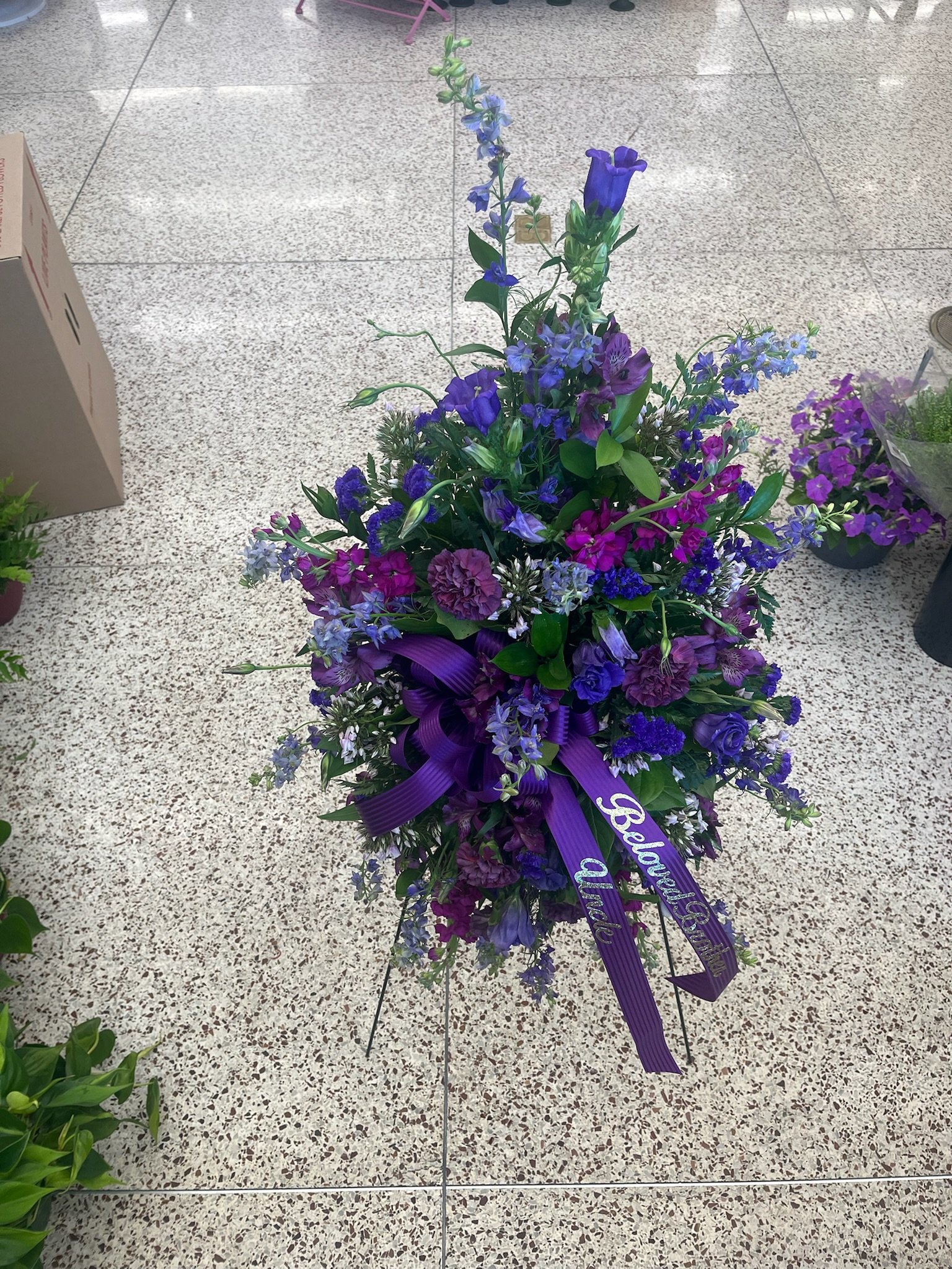 A colorful funeral flower arrangement with purple, pink, and blue flowers, decorated with purple ribbons that read "Peace" and "Alto," placed on a stand on a tile floor.