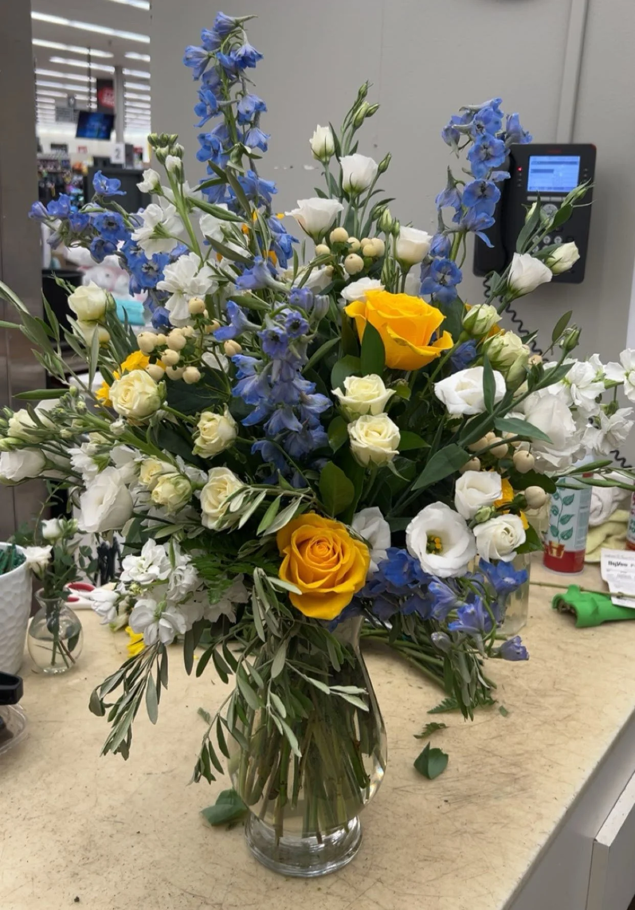 A colorful floral arrangement in a glass vase on a beige countertop in what appears to be a store or flower shop, featuring white, yellow, and blue flowers with green foliage.