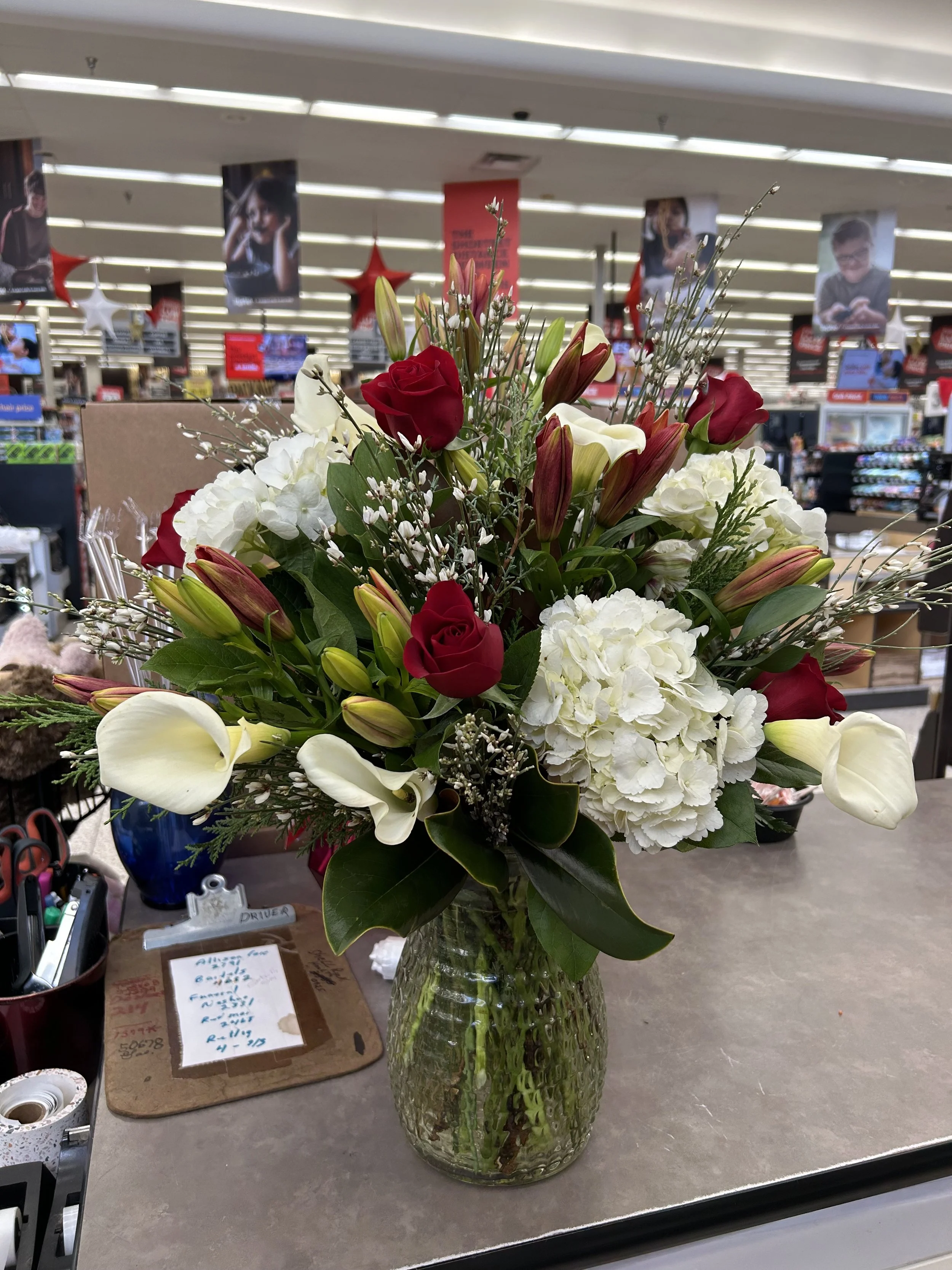 A floral arrangement in a textured clear glass vase, containing white hydrangeas, red roses, white calla lilies, pink lilies, and assorted greenery, placed on a store counter.