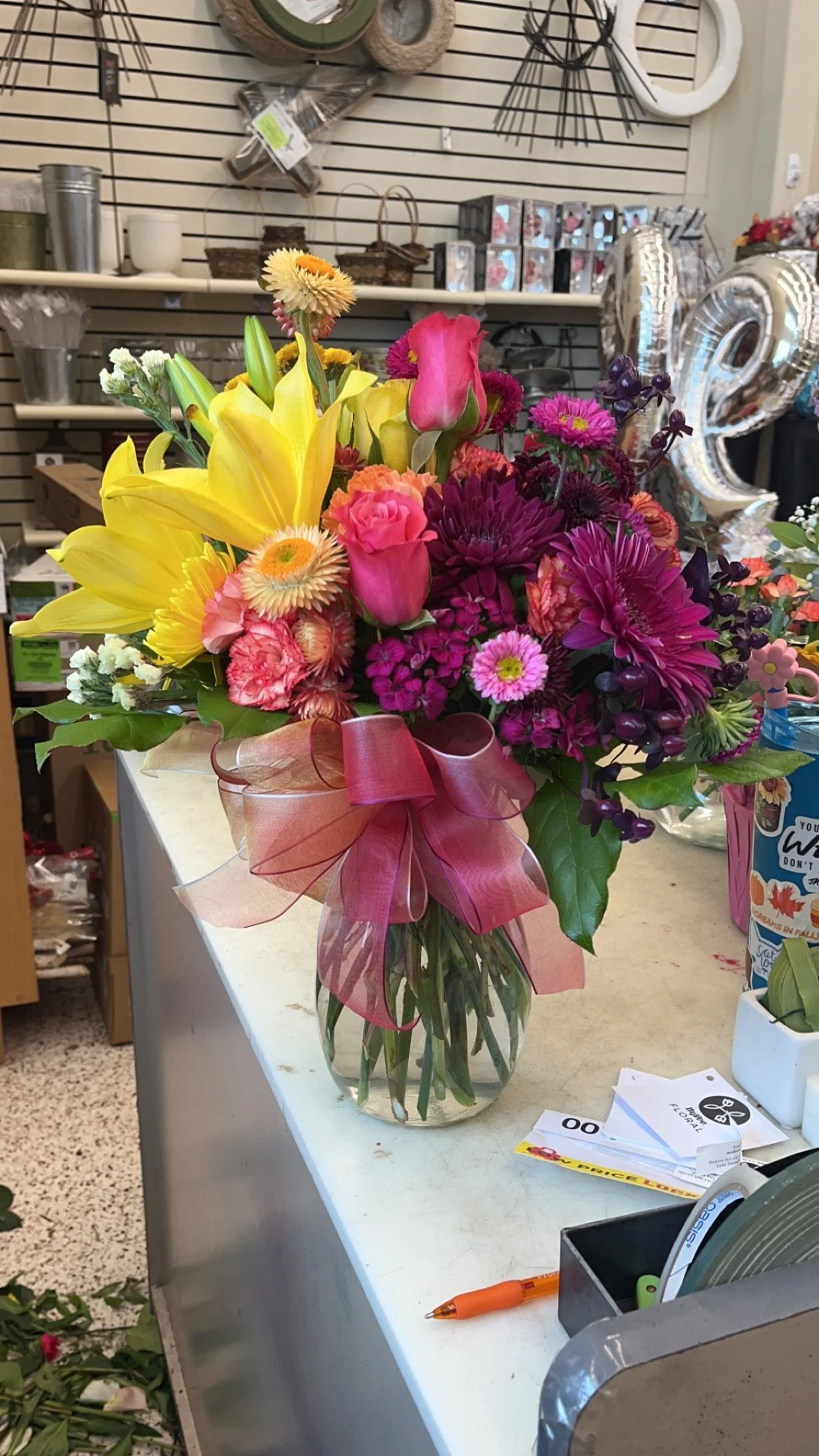 A colorful bouquet of flowers in a glass vase on a store counter, including yellow lilies, pink roses, purple daisies, and other mixed flowers, decorated with a pink ribbon.