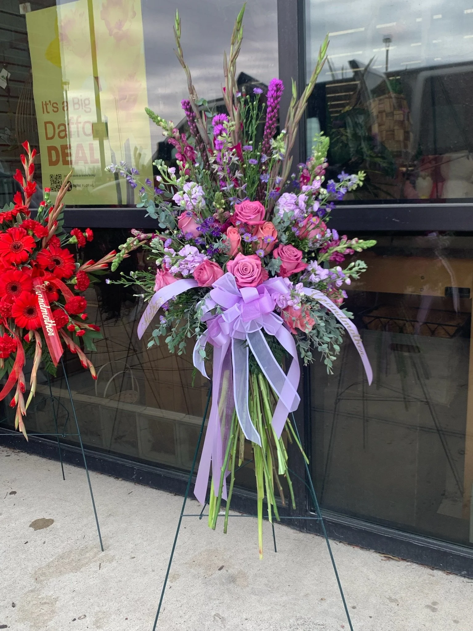 A large floral arrangement with pink roses, purple flowers, lavender, and greenery, decorated with a lavender ribbon, standing on a metal stand outside a building with glass windows.