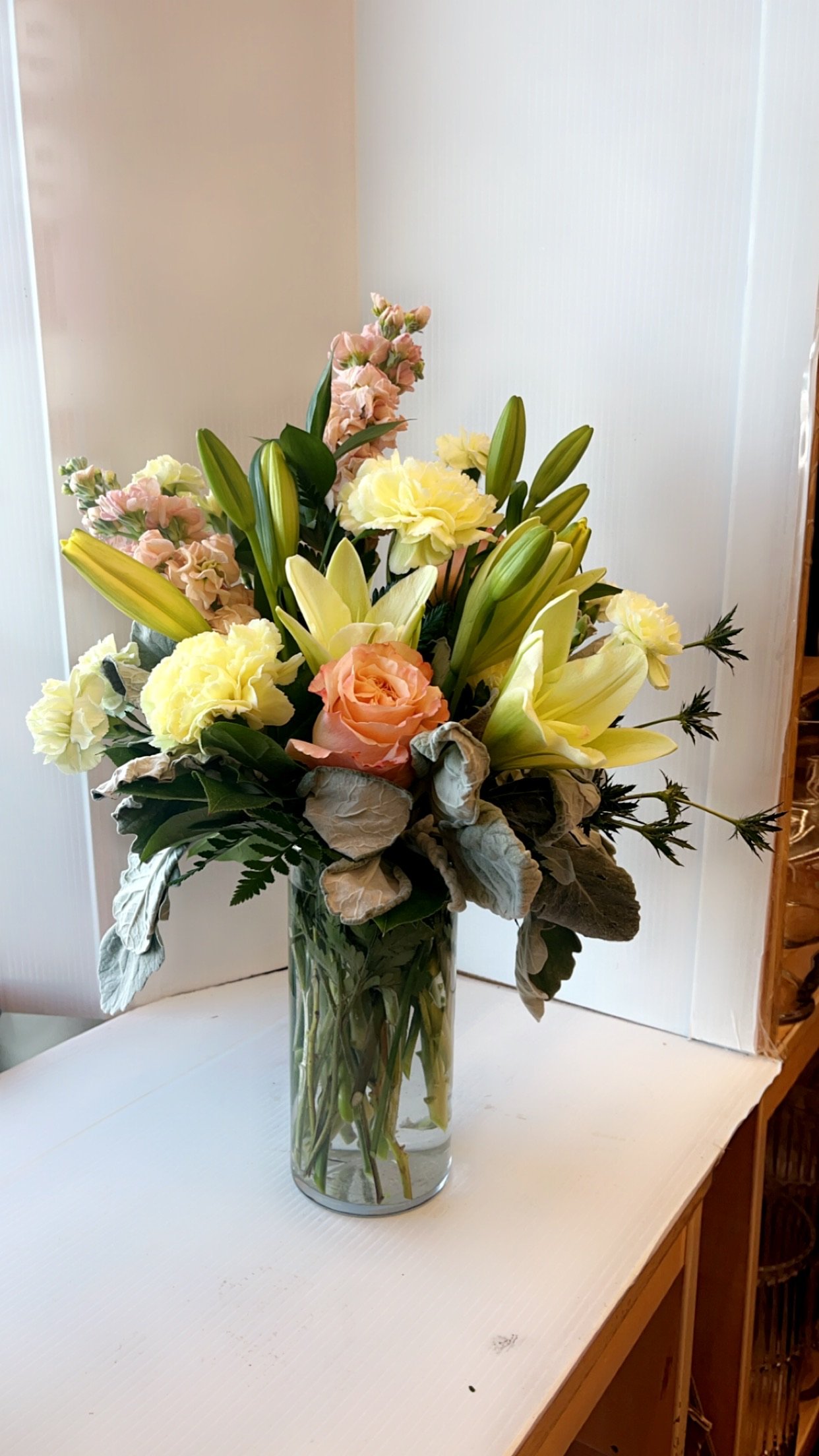A bouquet of mixed flowers, including lilies, roses, and carnations, in a clear glass vase on a white surface.