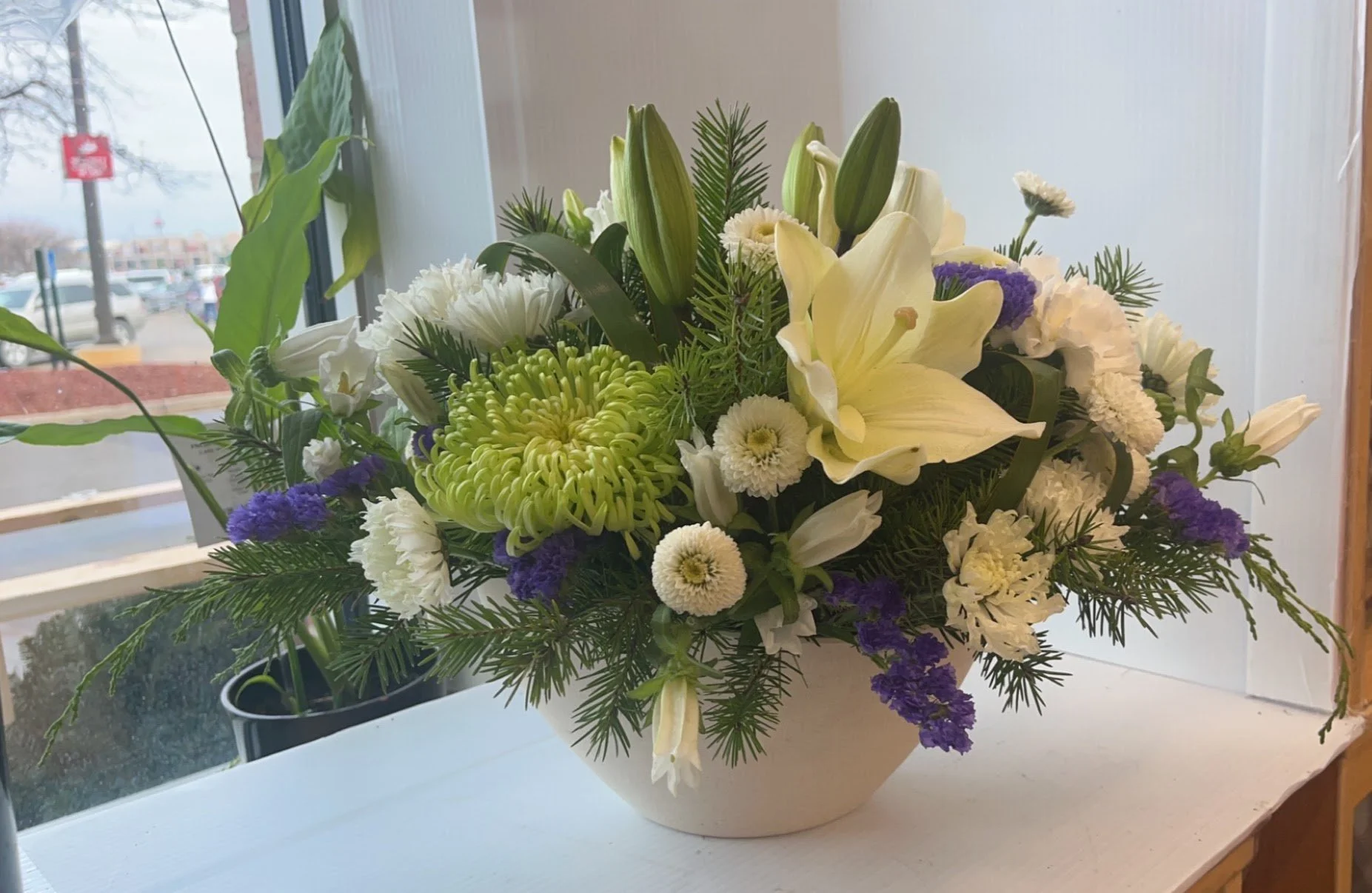 A floral arrangement with white lilies, chrysanthemums, and purple statice in a cream-colored bowl, placed on a white windowsill with a window showing a parking lot outside.