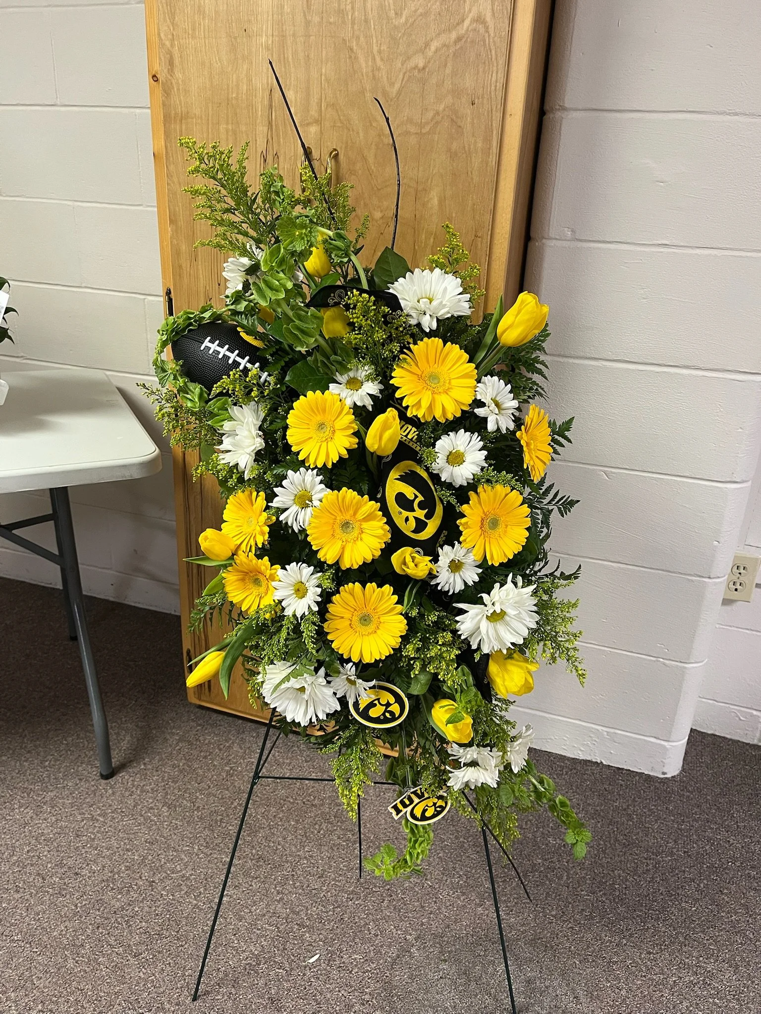 Yellow and white floral arrangement with Iowa Hawkeyes themed decorations, including a football and a badge, on a black metal stand in a room with white brick walls.