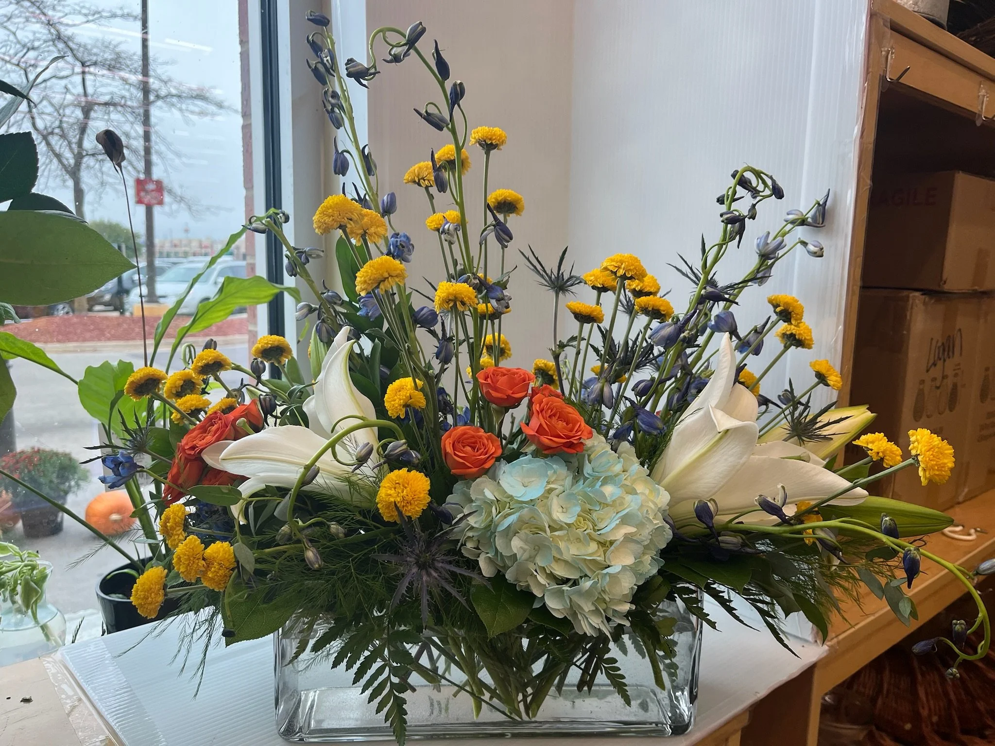 A floral arrangement with white lilies, orange roses, blue irises, and yellow daisies in a clear glass vase on a table near a window.