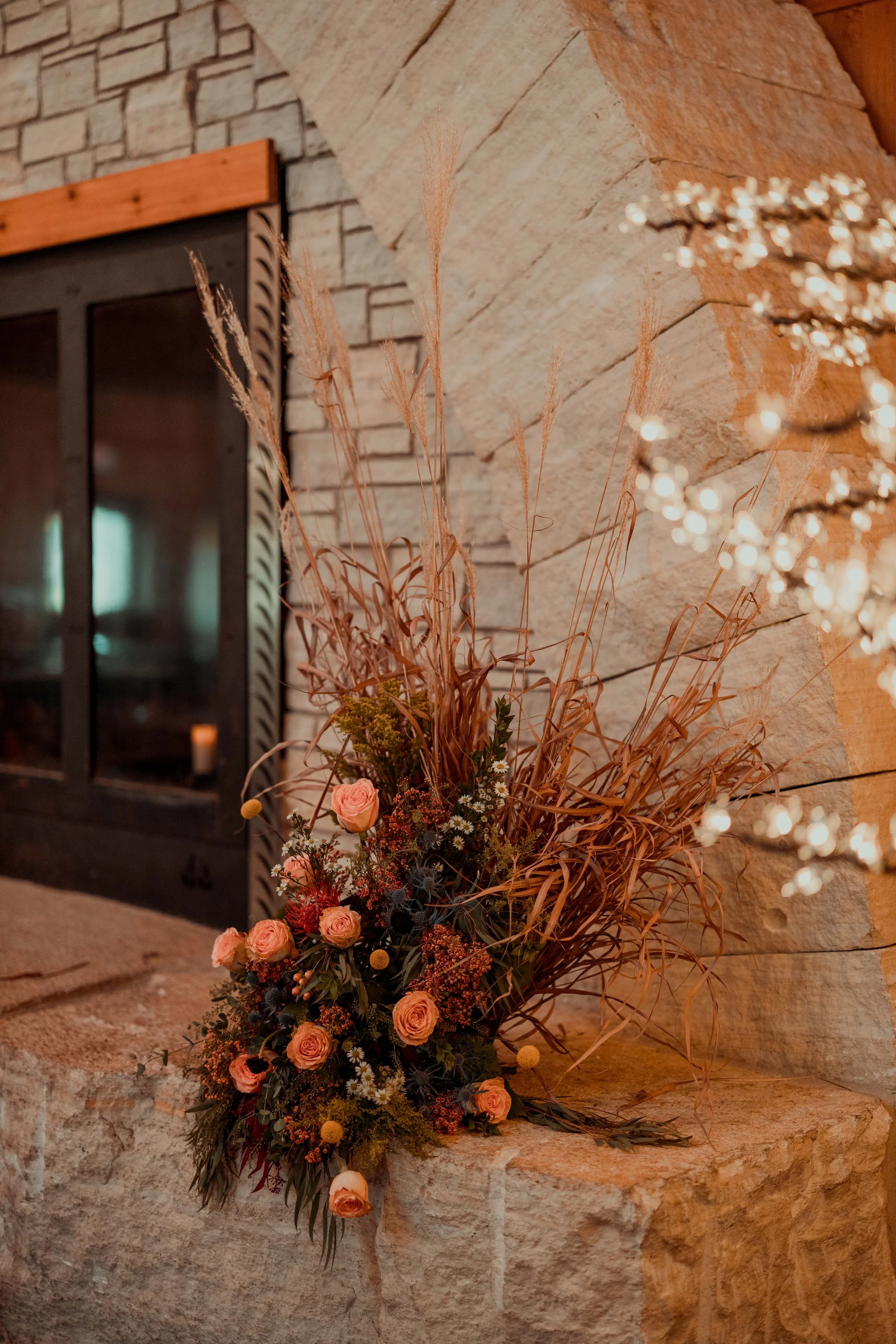 A floral arrangement with pink roses, dried grasses, and small white and yellow flowers, placed on a stone mantel in front of a stone and wood fireplace.