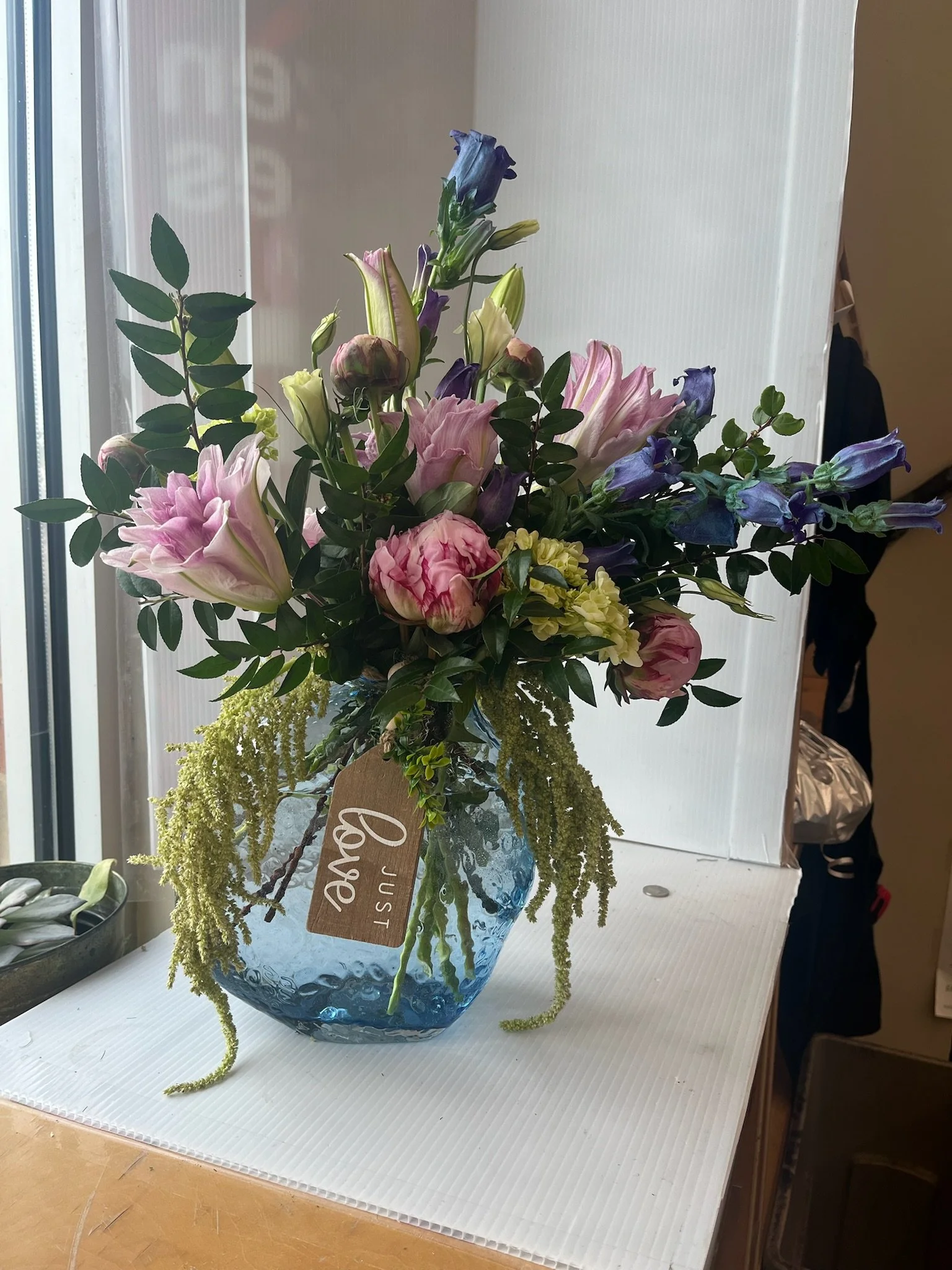 A bouquet of pink, purple, and white flowers in a blue glass vase with a wooden tag that says 'Just Love' on it.