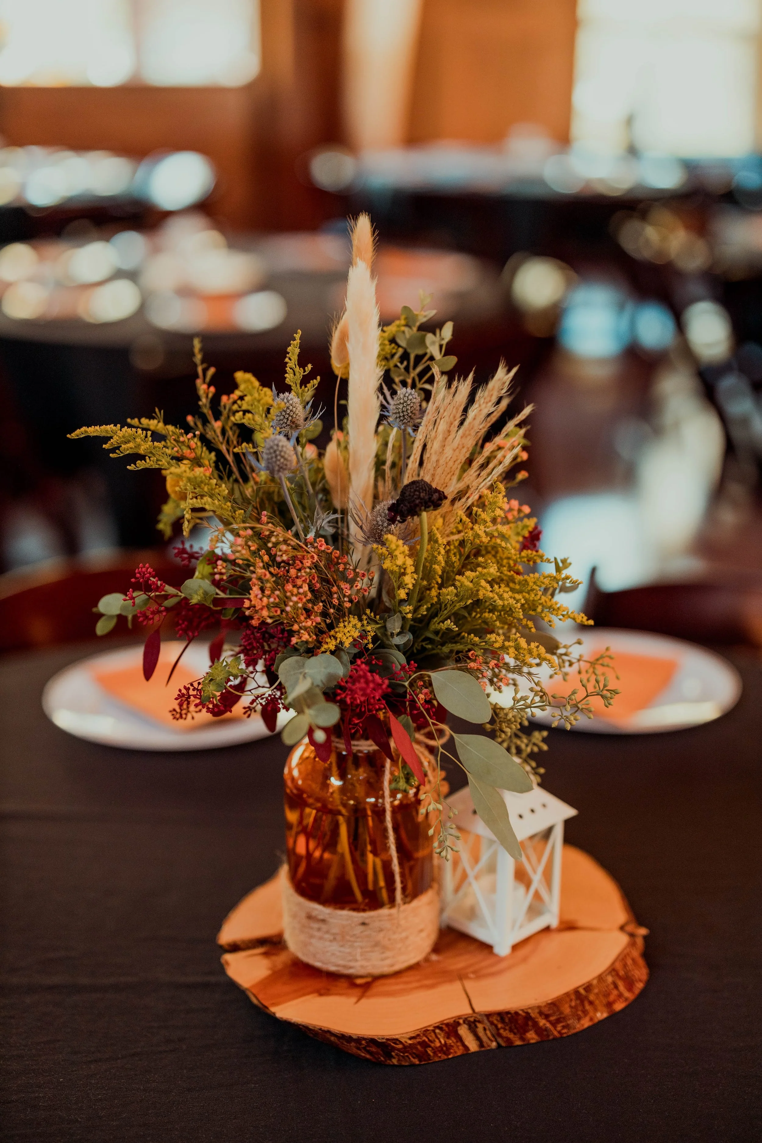 A floral centerpiece with various dried and fresh flowers and greenery in a amber glass jar on a wooden slab, with a small white lantern beside it on a black table.