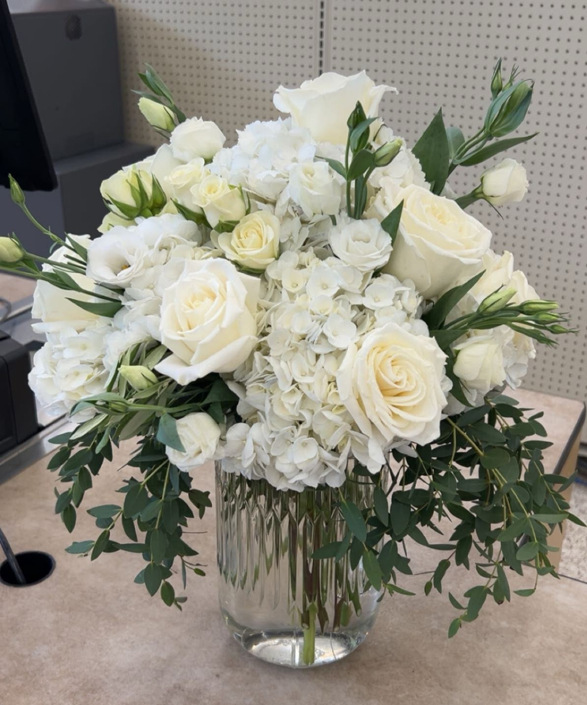 A large bouquet of white roses, hydrangeas, and other white flowers in a tall, clear, patterned glass vase on a beige surface.