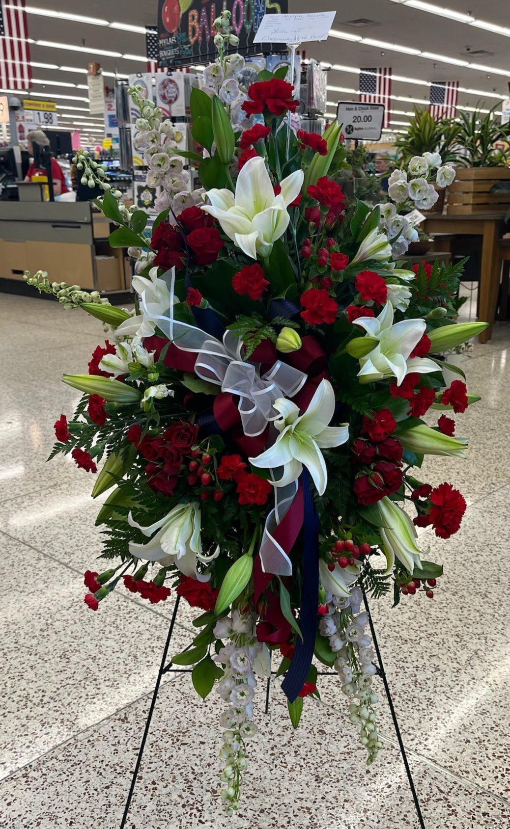 A funeral flower arrangement featuring white lilies, red roses, white orchids, and greenery, displayed on a black stand in a store aisle.