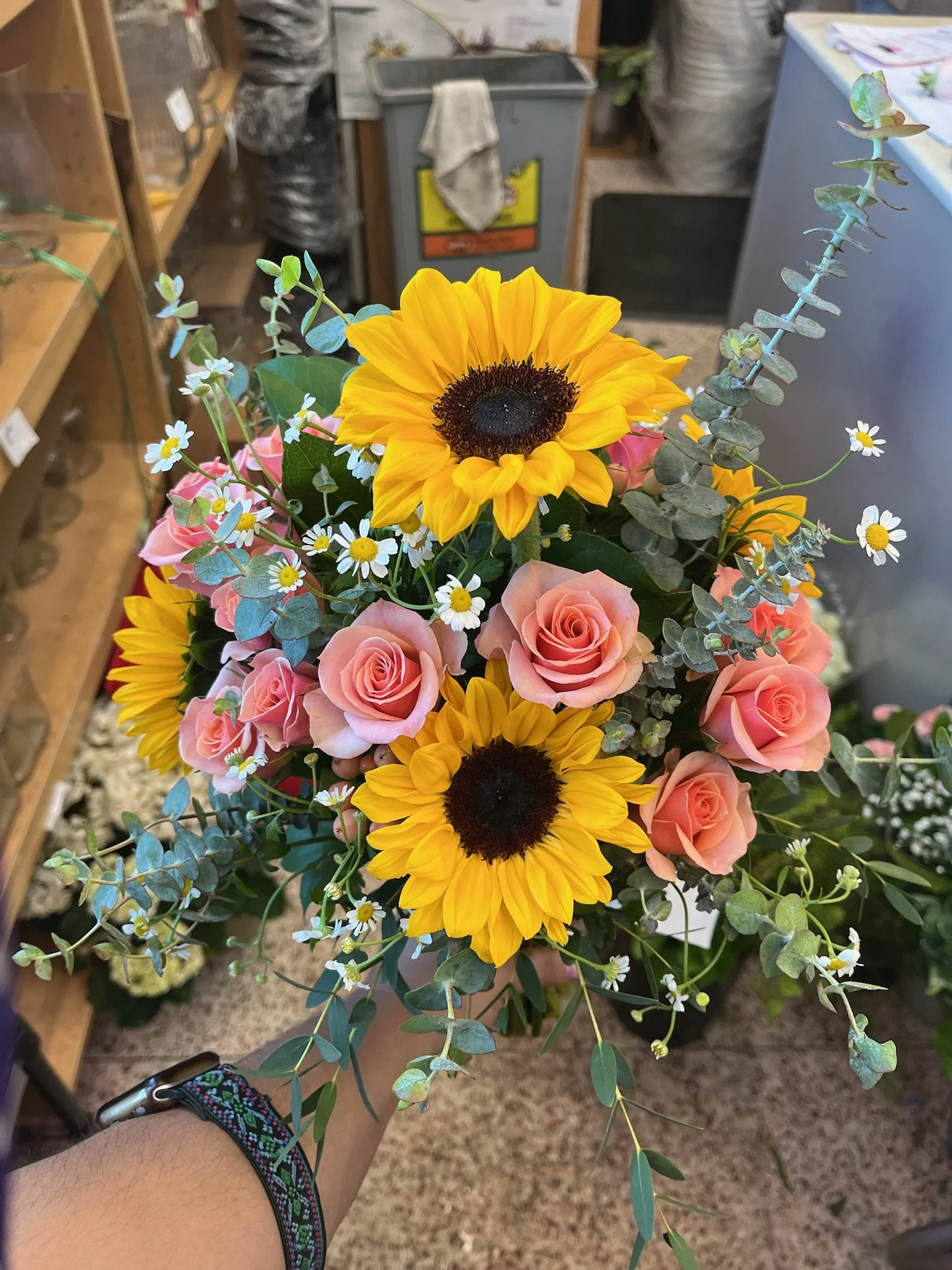 Bouquet of flowers with yellow sunflowers, pink roses, small white daisies, and greenery, held by a person wearing a black watch with a colorful strap.