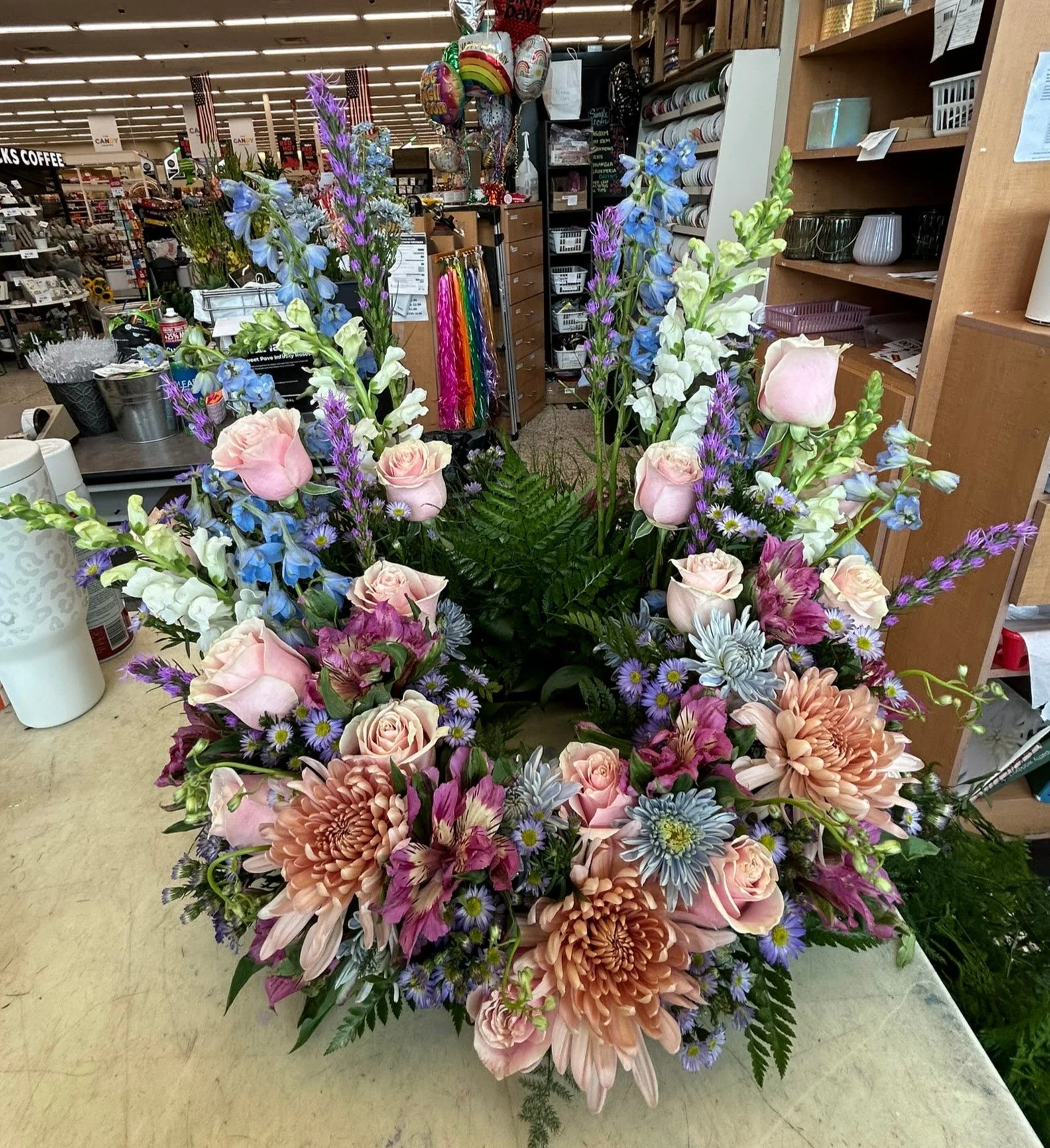 A colorful flower arrangement featuring pink roses, peach chrysanthemums, blue delphiniums, purple statice, and various green foliage, placed on a countertop inside a store.