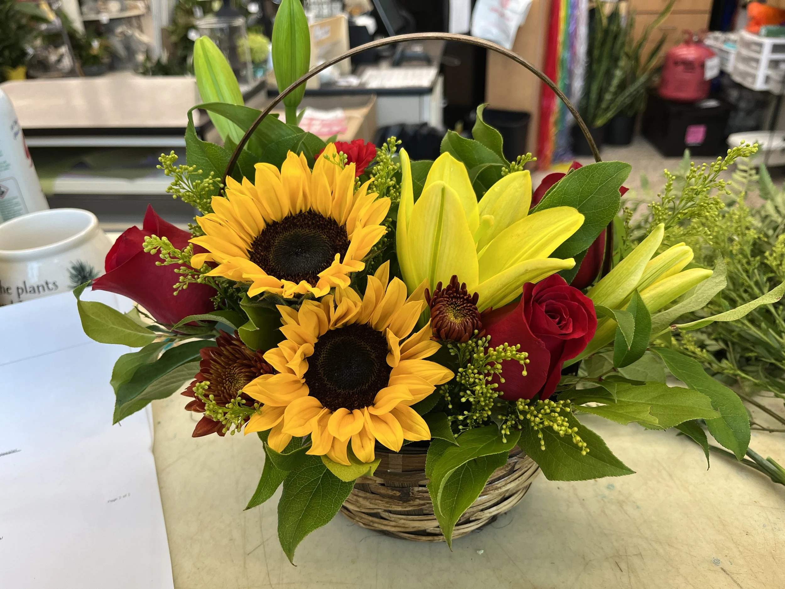 A basket of yellow sunflowers, red roses, and yellow lilies with greenery inside a store.