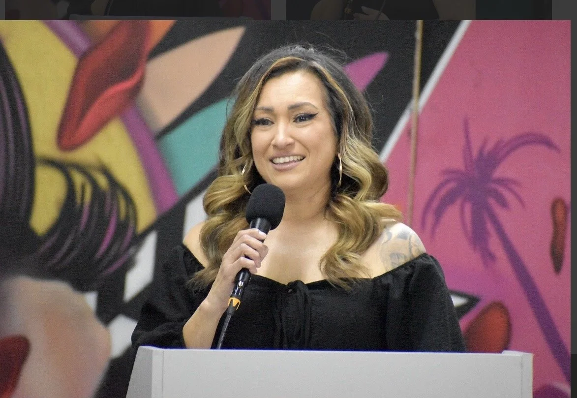 A woman with wavy, shoulder-length hair speaking into a microphone at a podium, with a colorful mural featuring tropical elements in the background.