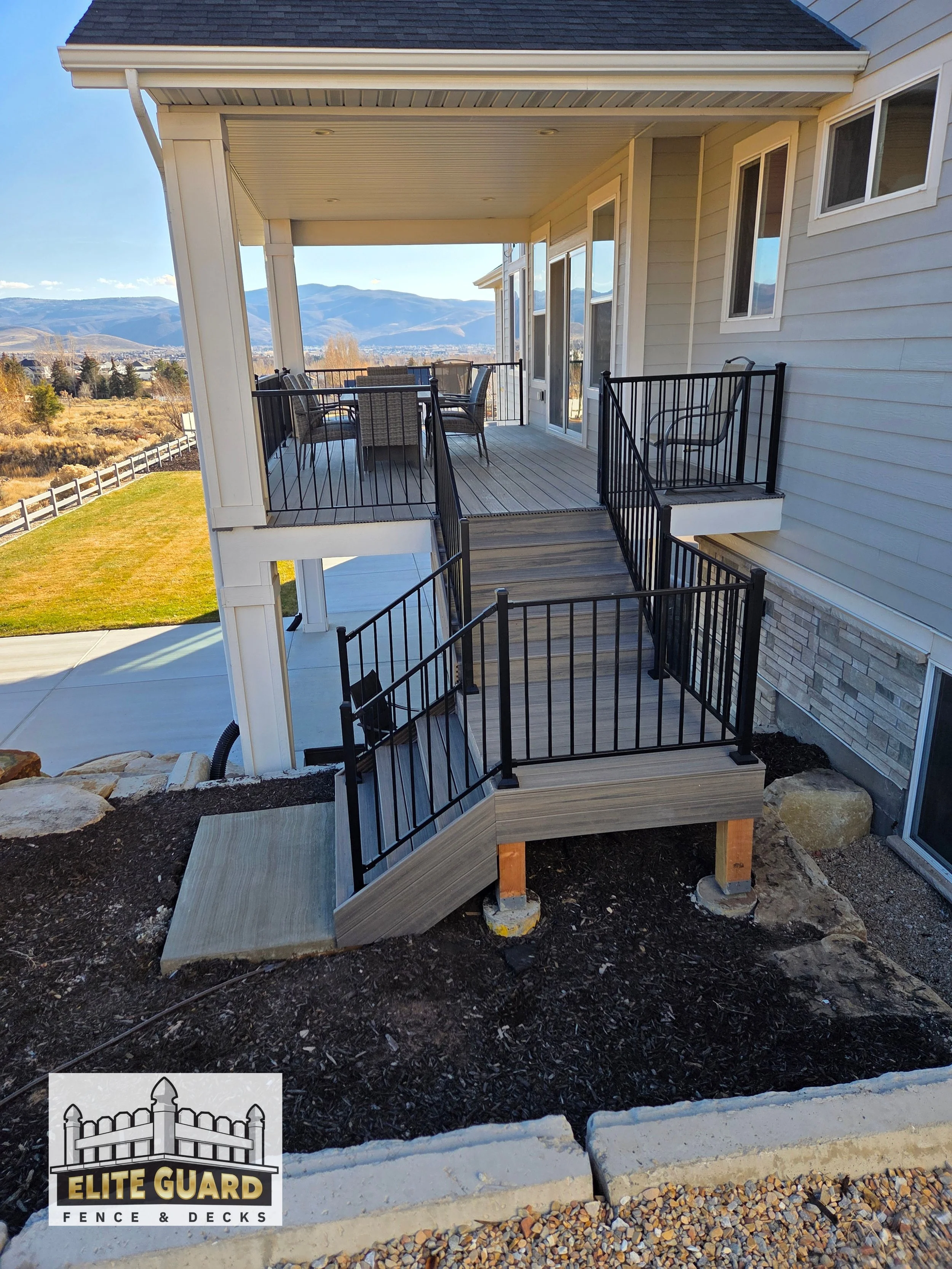 Backyard deck with black metal railings, accessed by a staircase, attached to a house with beige siding, overlooking a landscape with mountains in the distance under a clear blue sky in Payson, Utah.