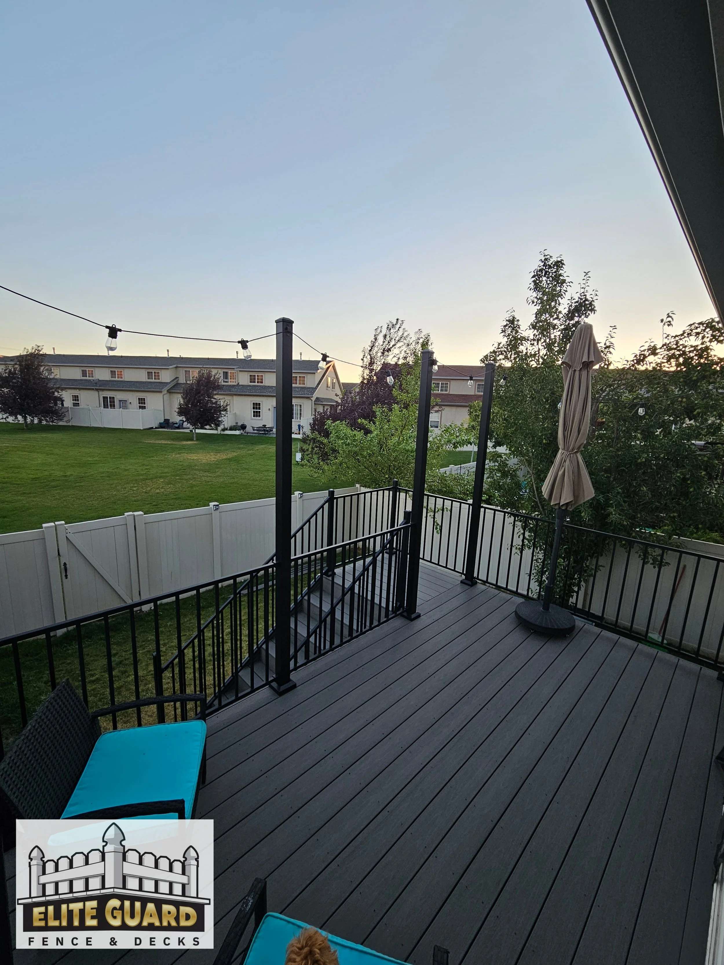 Photo of a backyard deck with a black railing, outdoor chair with teal cushion, and a closed patio umbrella. The background shows a grassy yard, trees, and a row of neighboring houses against a clear sky during sunset or dusk in Salem, Utah.